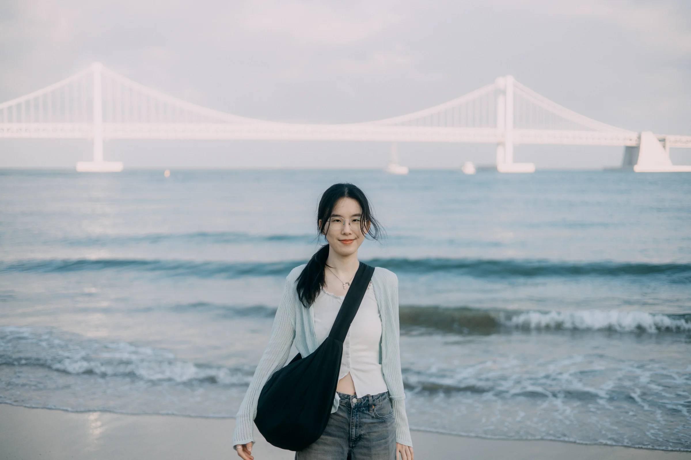 A person stands beside gentle waves beneath a pastel sky, with a bridge across the horizon.