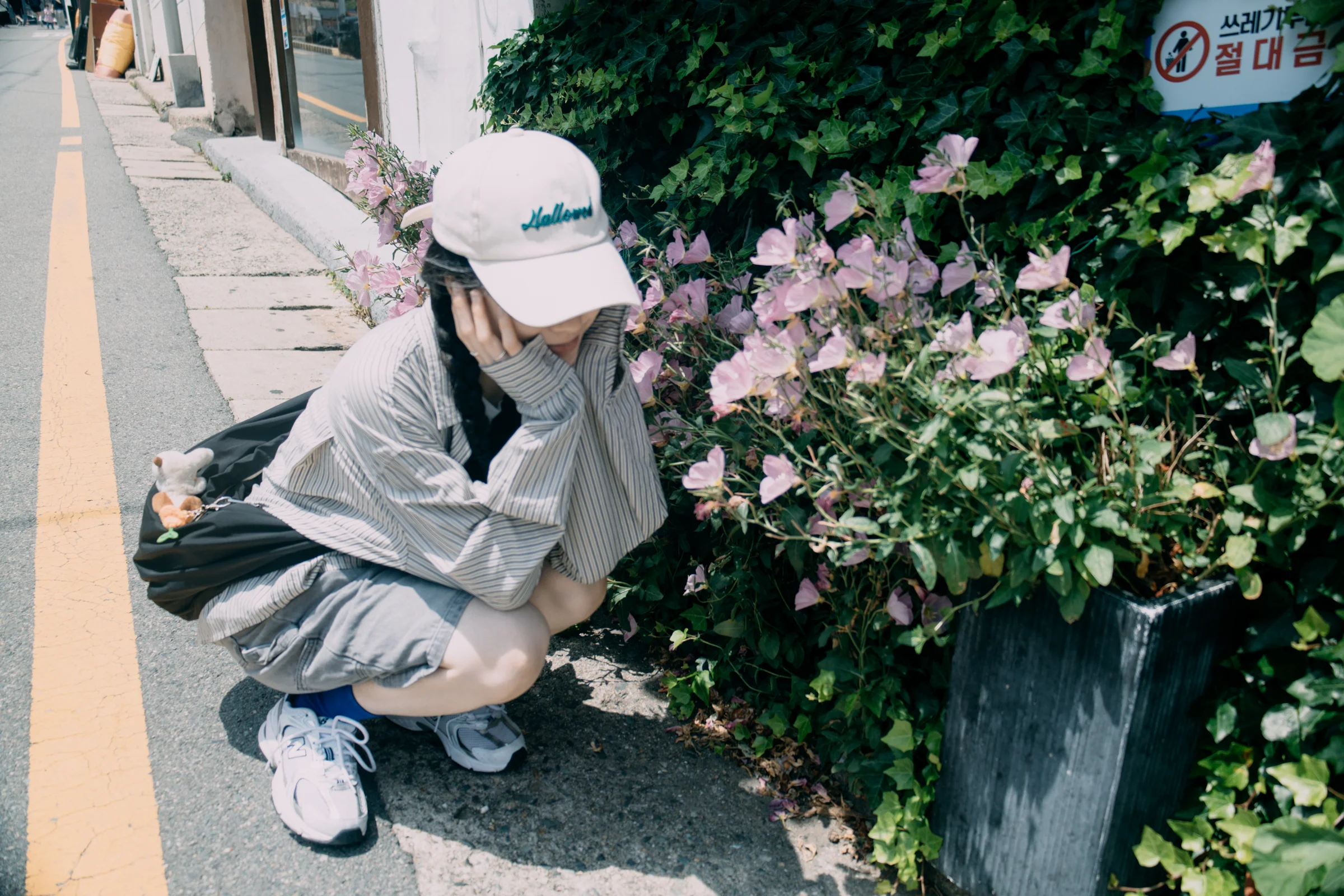 A person crouches beside pink hydrangeas along a sunlit street.