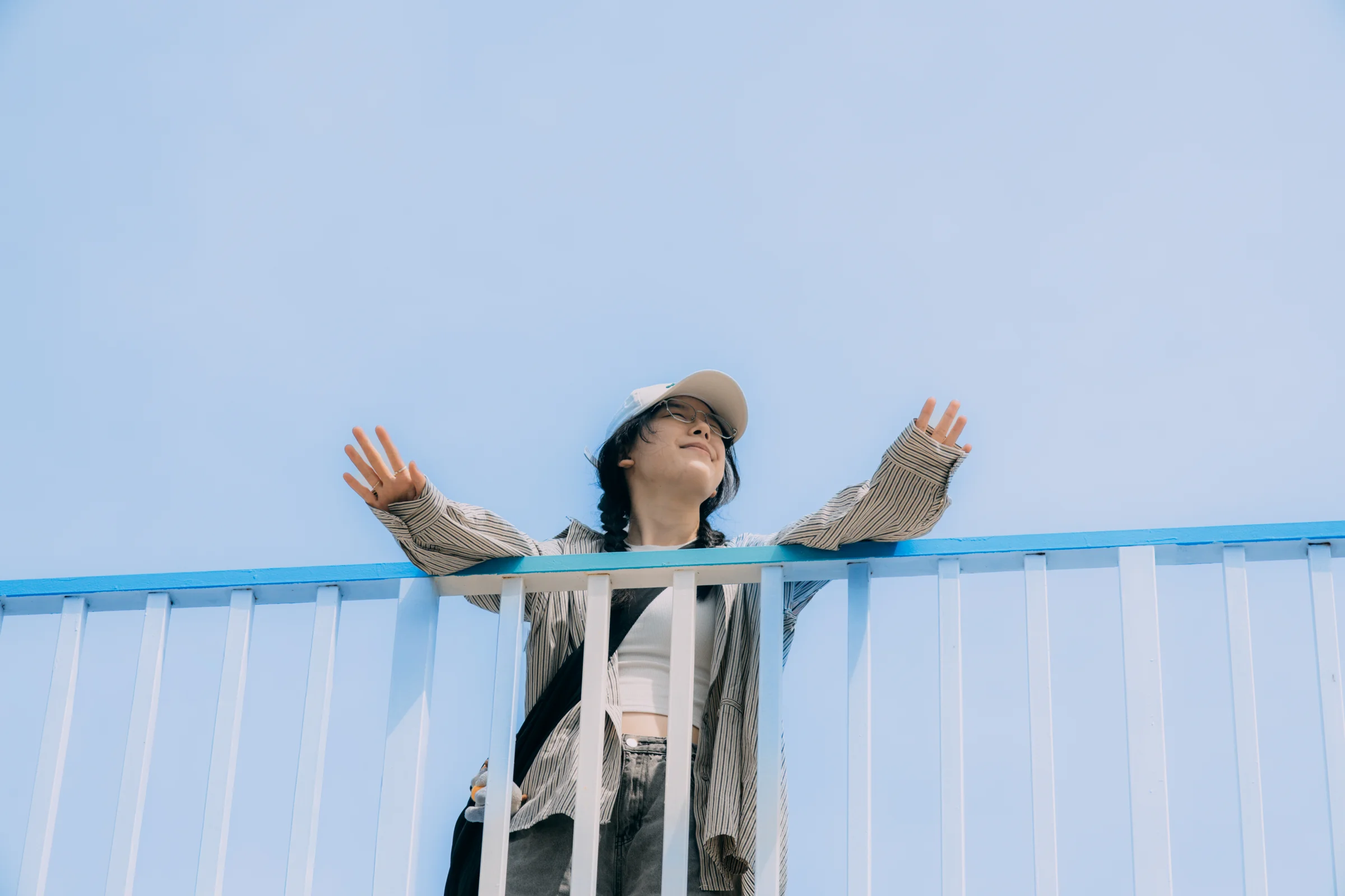 A person stretches their arms above a blue and white railing against a clear sky.