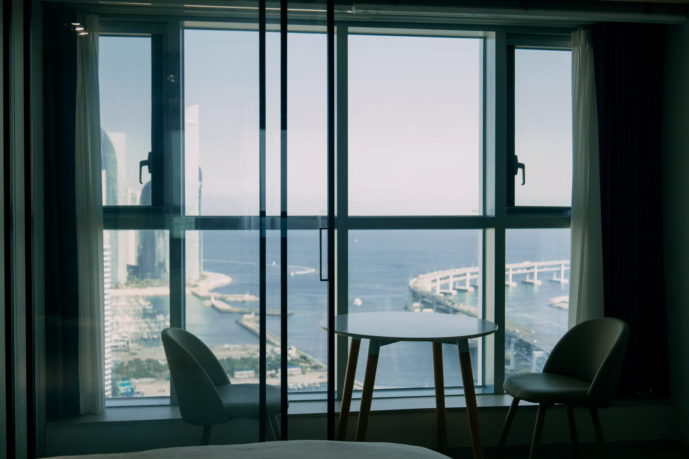 A table and chairs sit in silhouette before tall windows overlooking the coast.