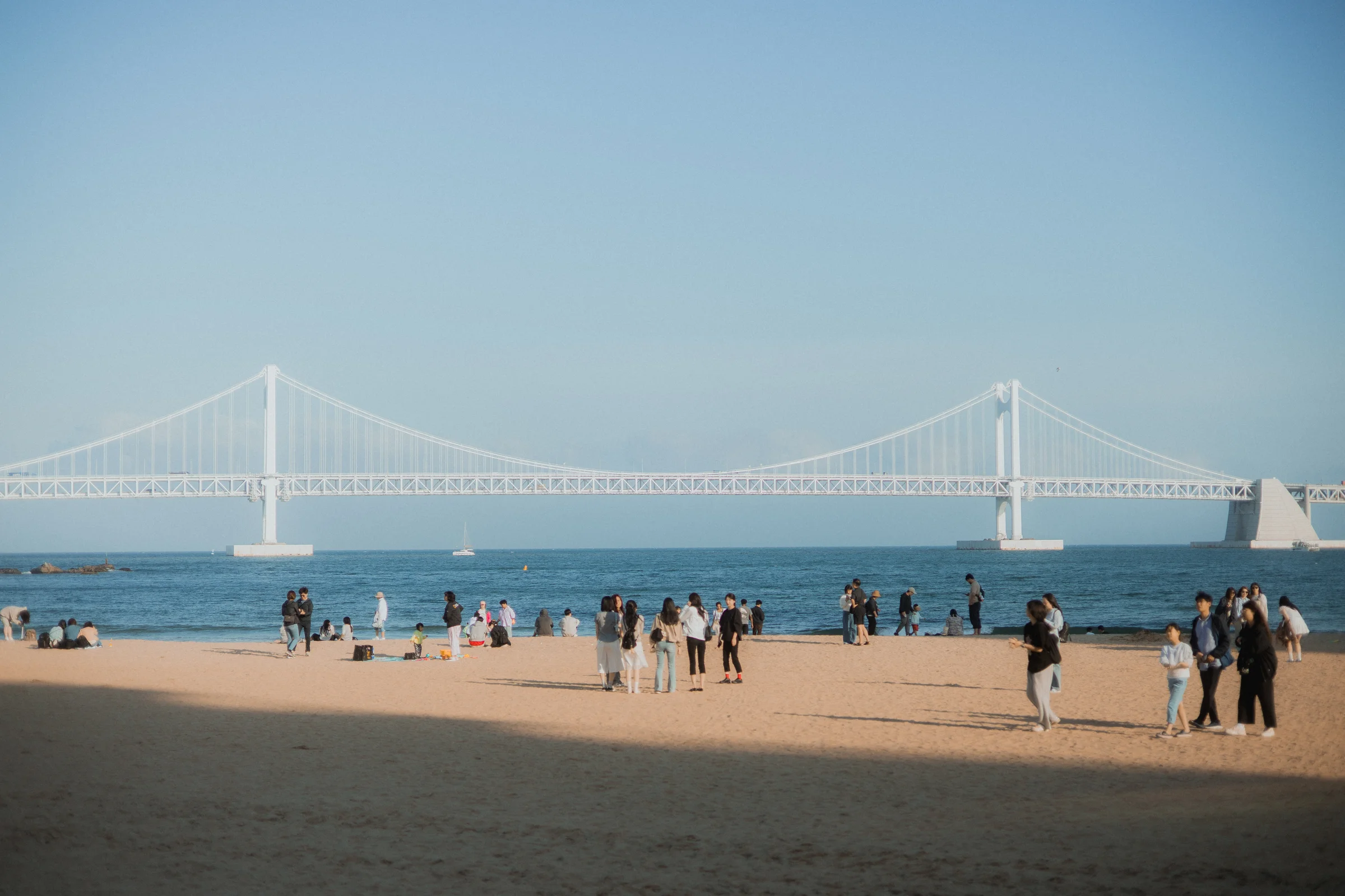 People gather on a sandy beach beneath a clear sky, with the Gwangan Bridge bridge stretching across the sea.