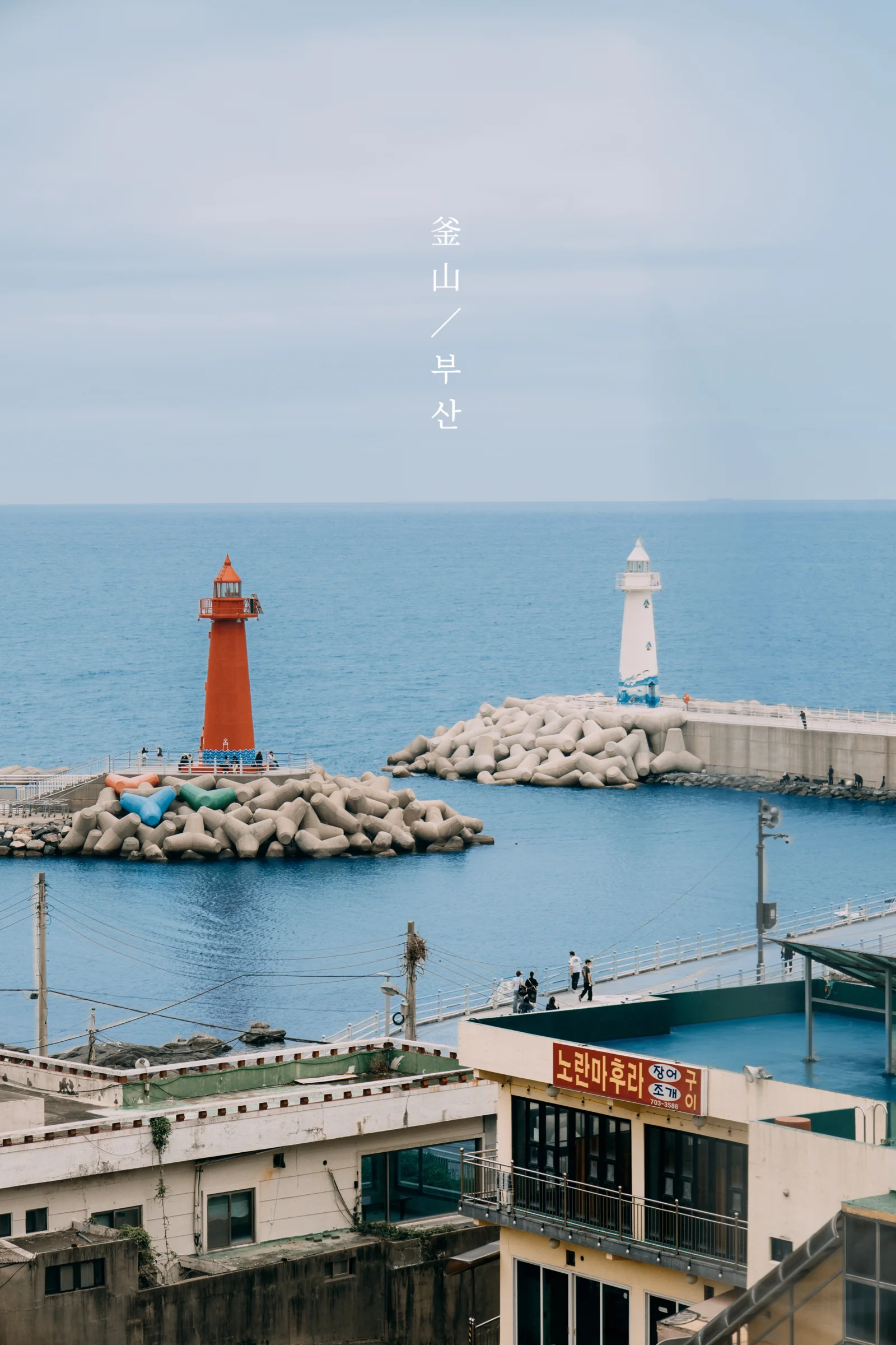 Red and white lighthouses stand on breakwaters beyond waterfront rooftops and blue harbor water.