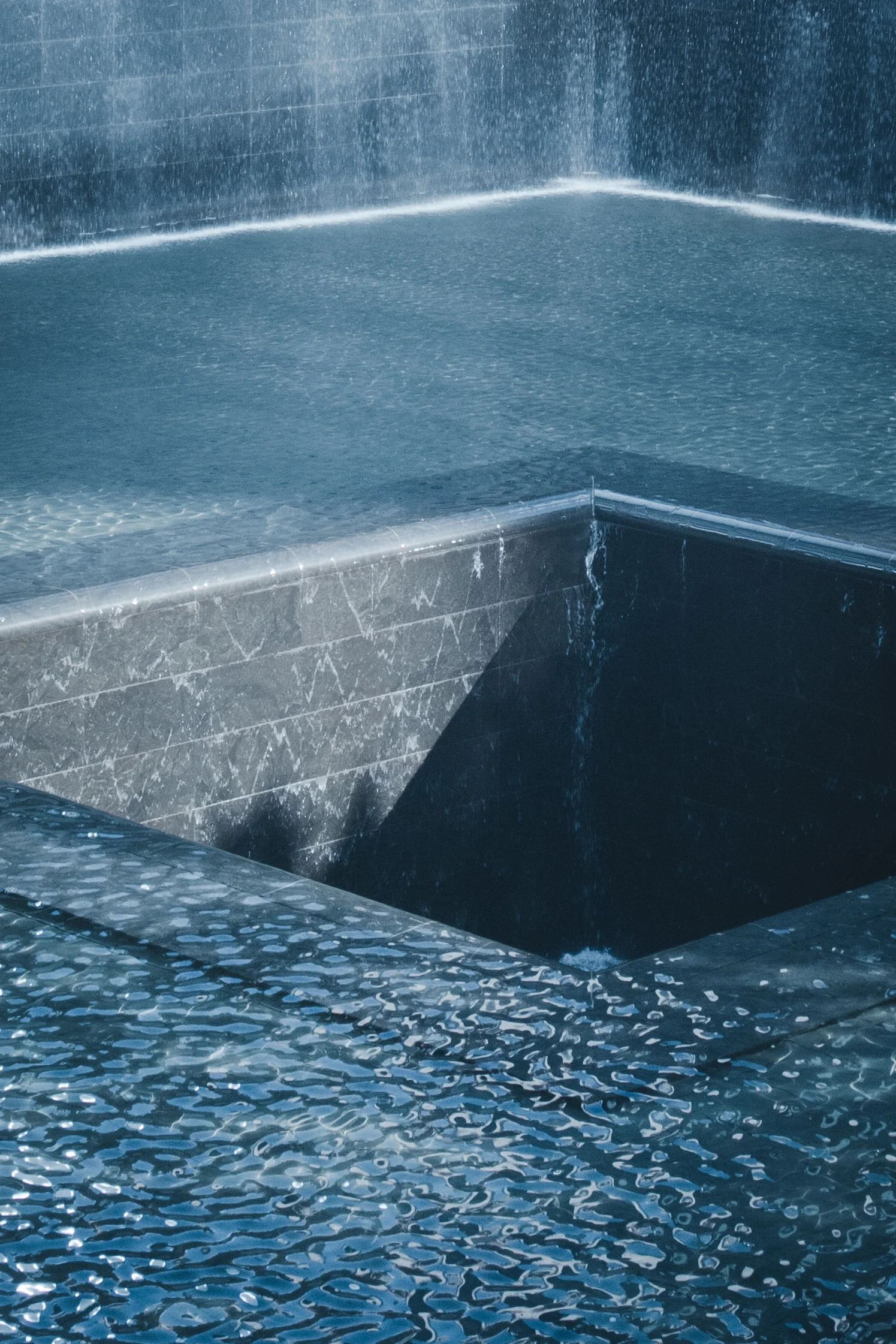 An angled view of a 911 memorial pool with rippling blue water flowing into a central opening.