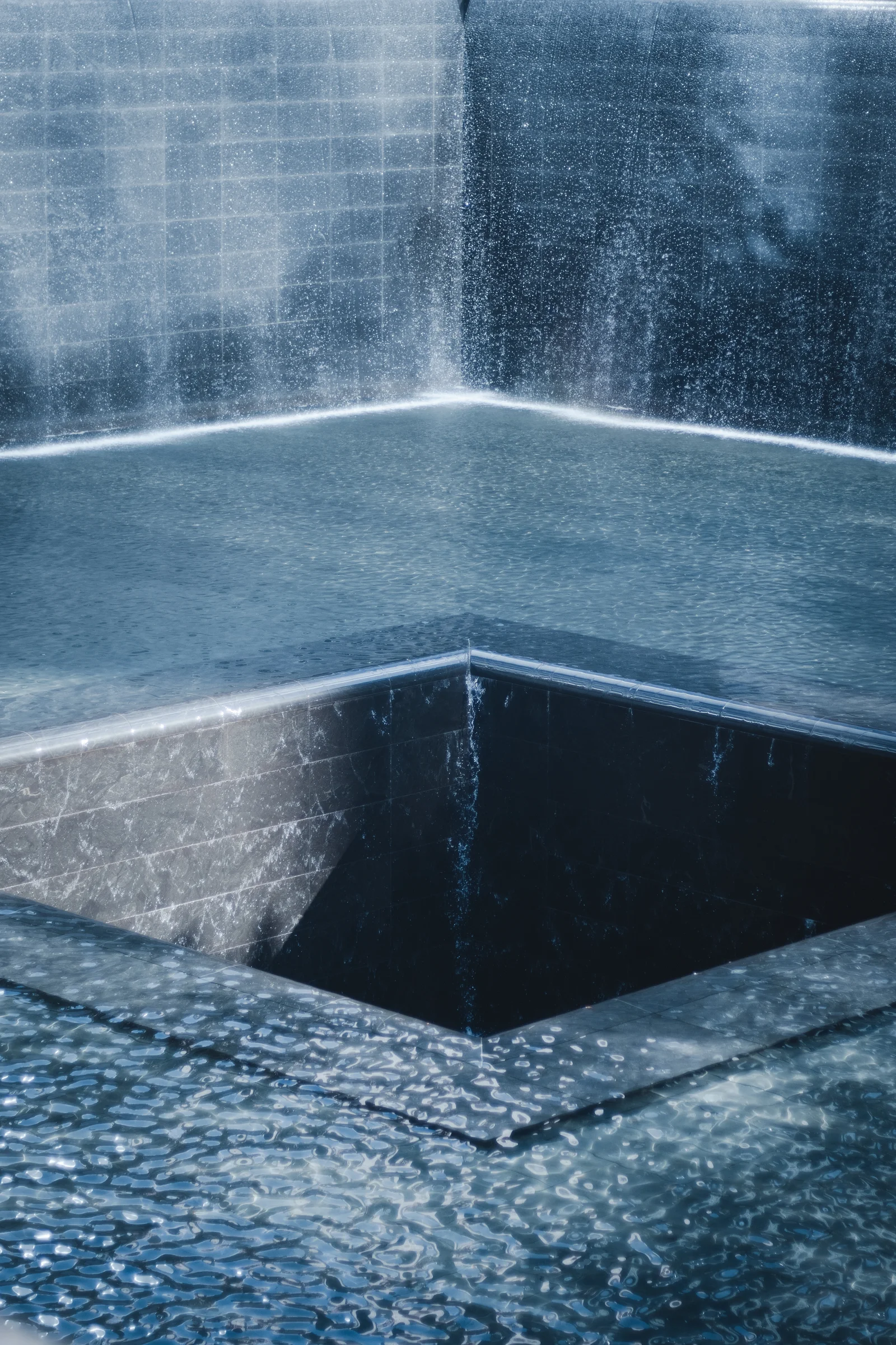 Sunlight catches the water cascading around a square void in a memorial pool.