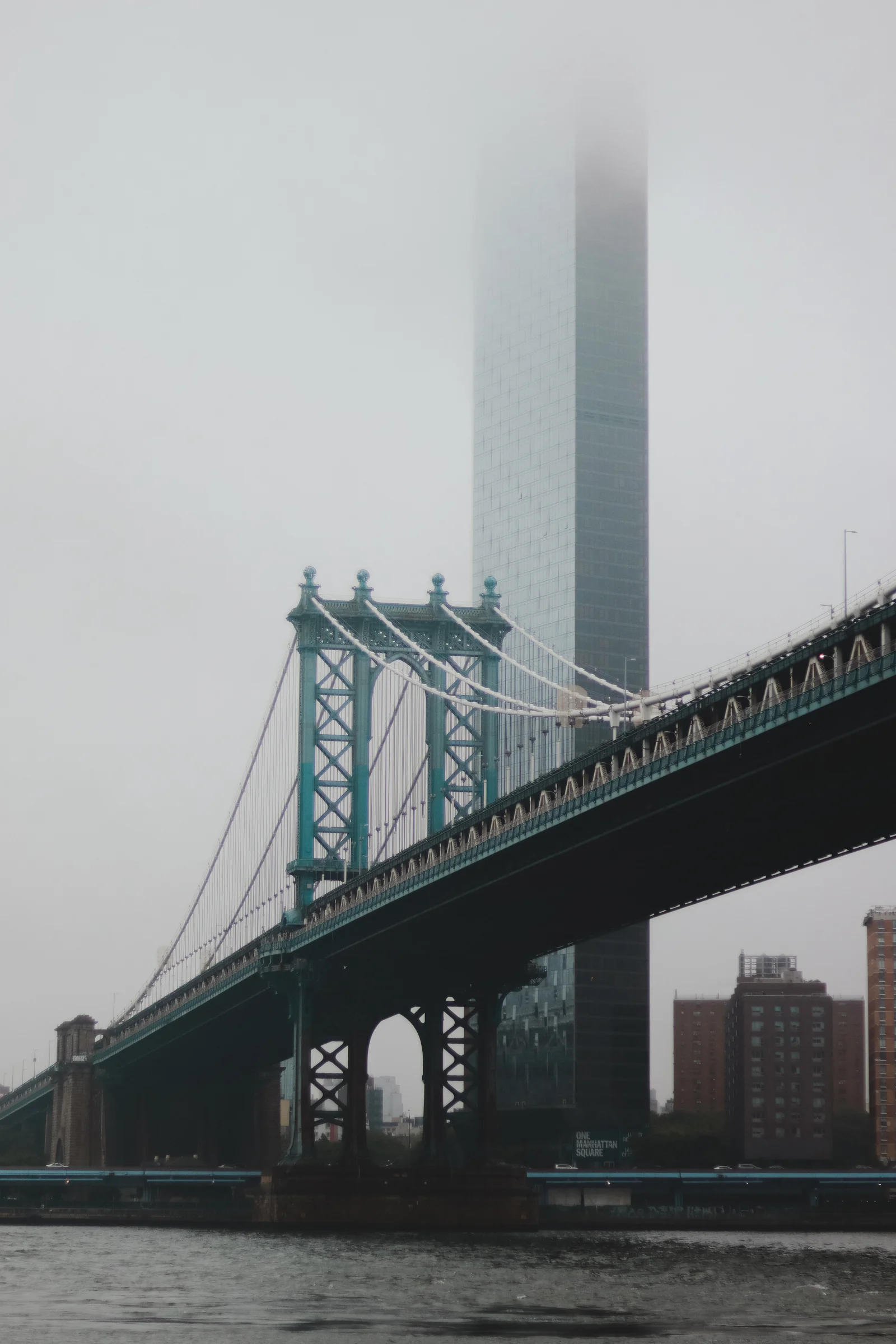 A steel suspension bridge crosses in front of a skyscraper disappearing into low clouds.