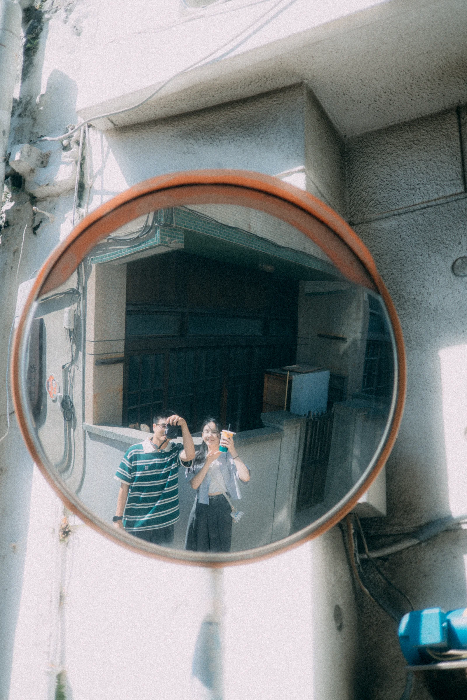 Two people appear in a round roadside mirror mounted beside a building.