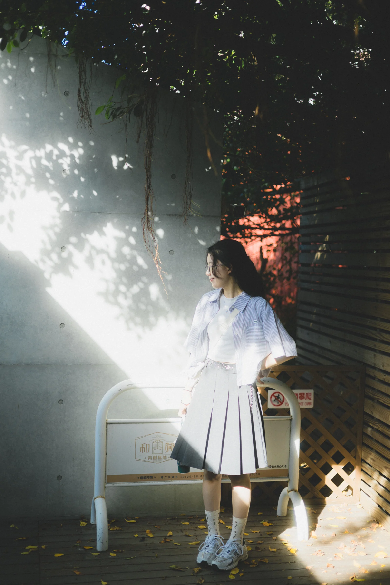 A person in a pale outfit stands beside a chair in dappled light beneath trees.