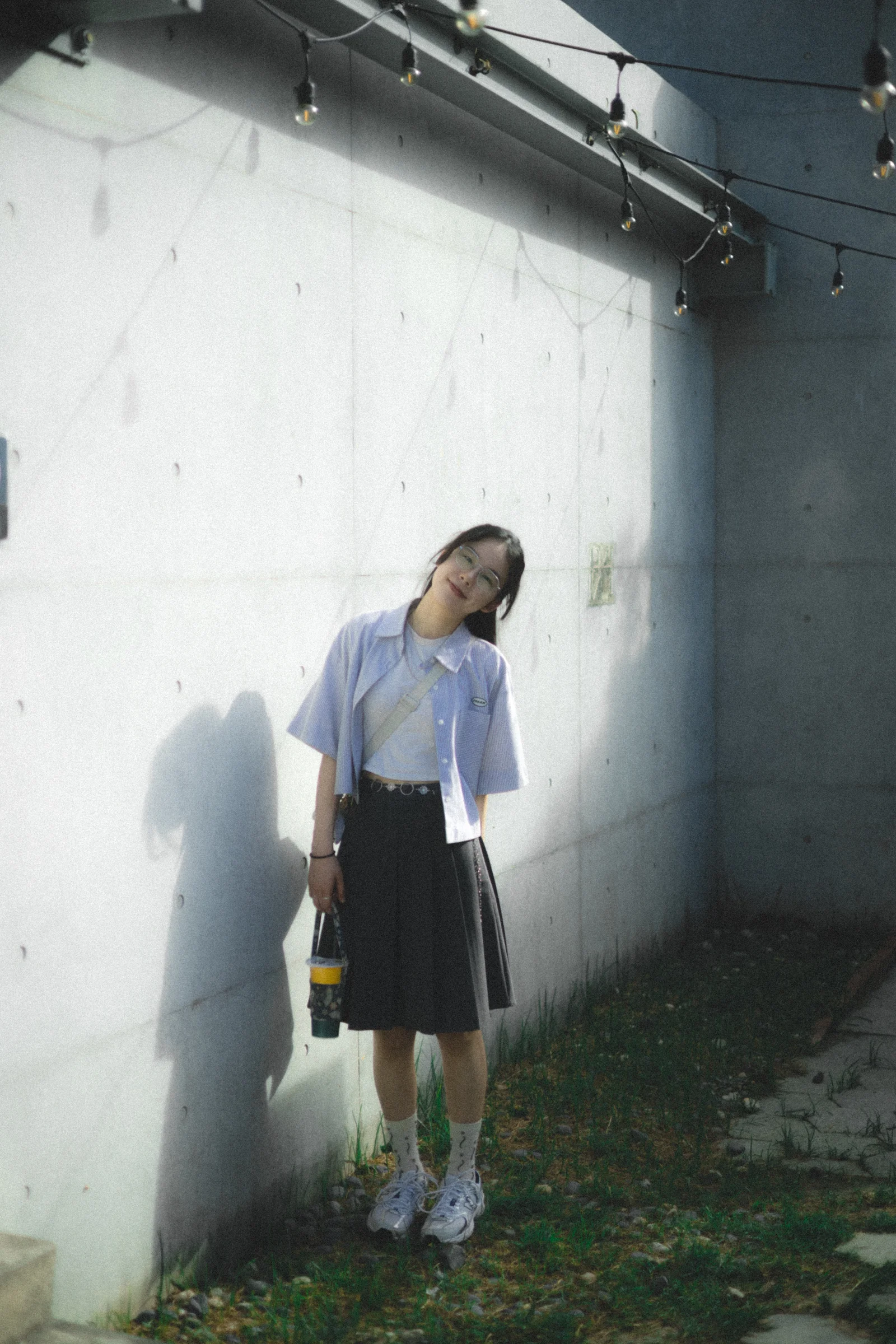 A person looks down while standing beside a white wall in a patch of sunlight.