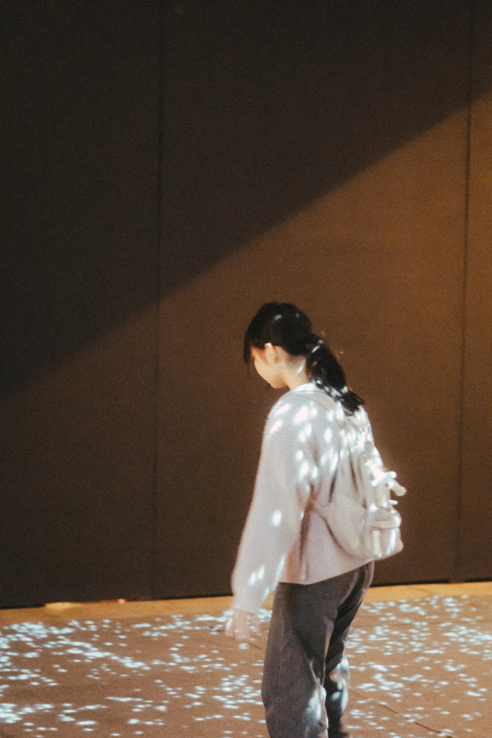 A person walks beside a dark wall as warm light falls across their pale shirt.