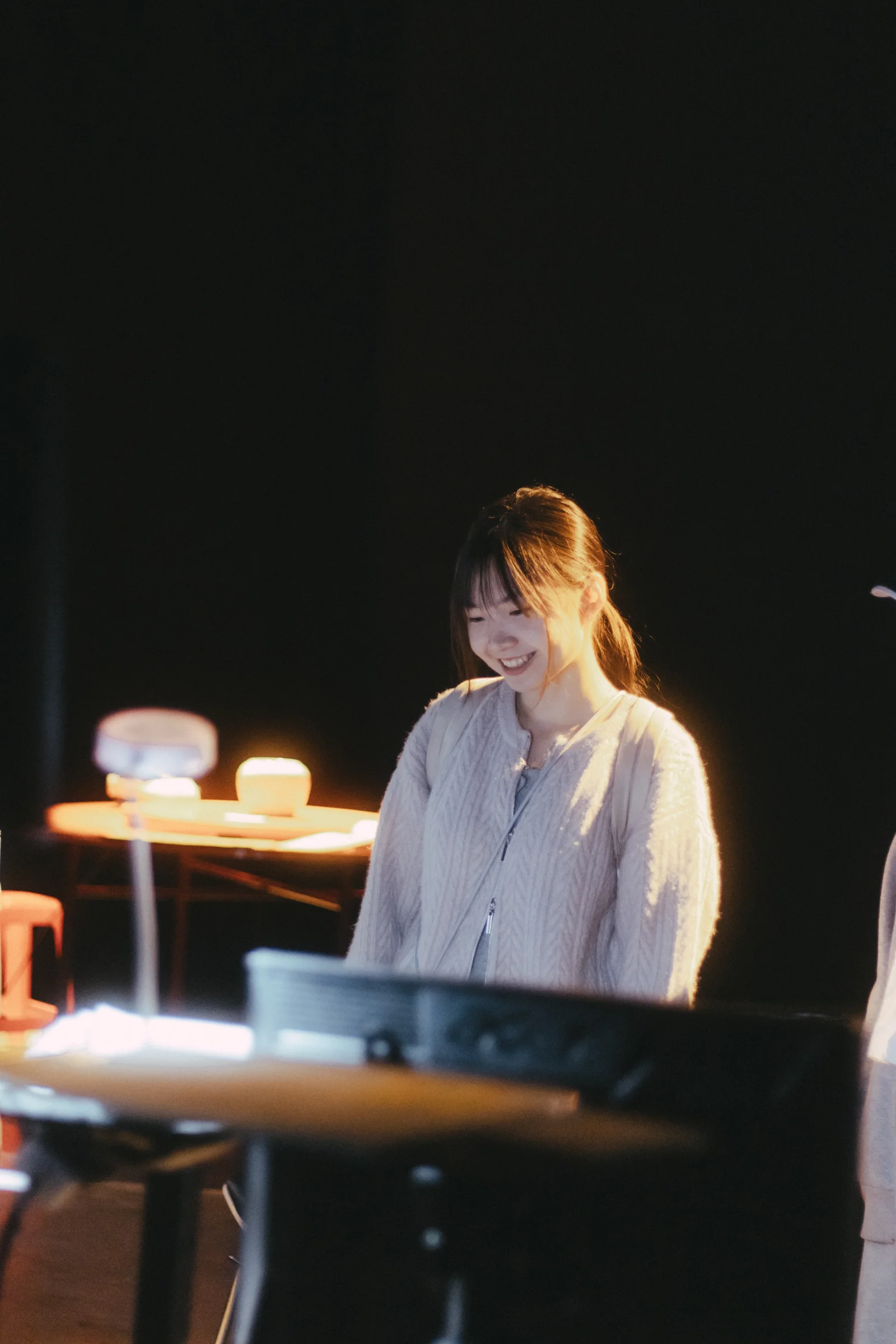 A seated person in a pale shirt faces a desk in a dark performance space.