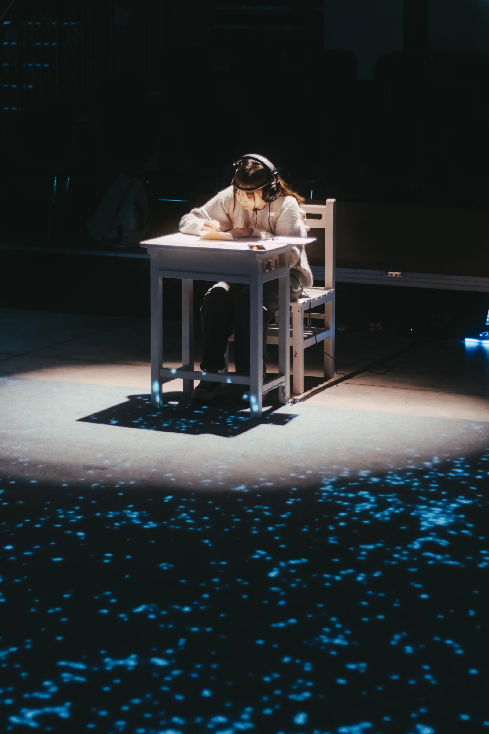 A performer sits at a desk illuminated by a spotlight on a dark stage.