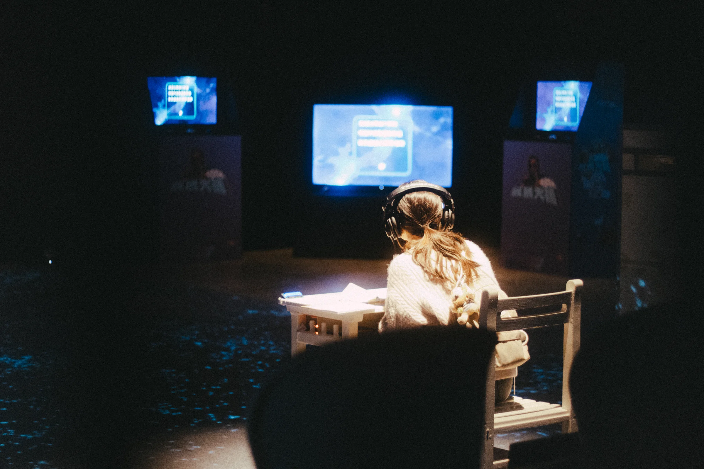 A performer sits at a lit desk facing glowing screens across a dark stage.