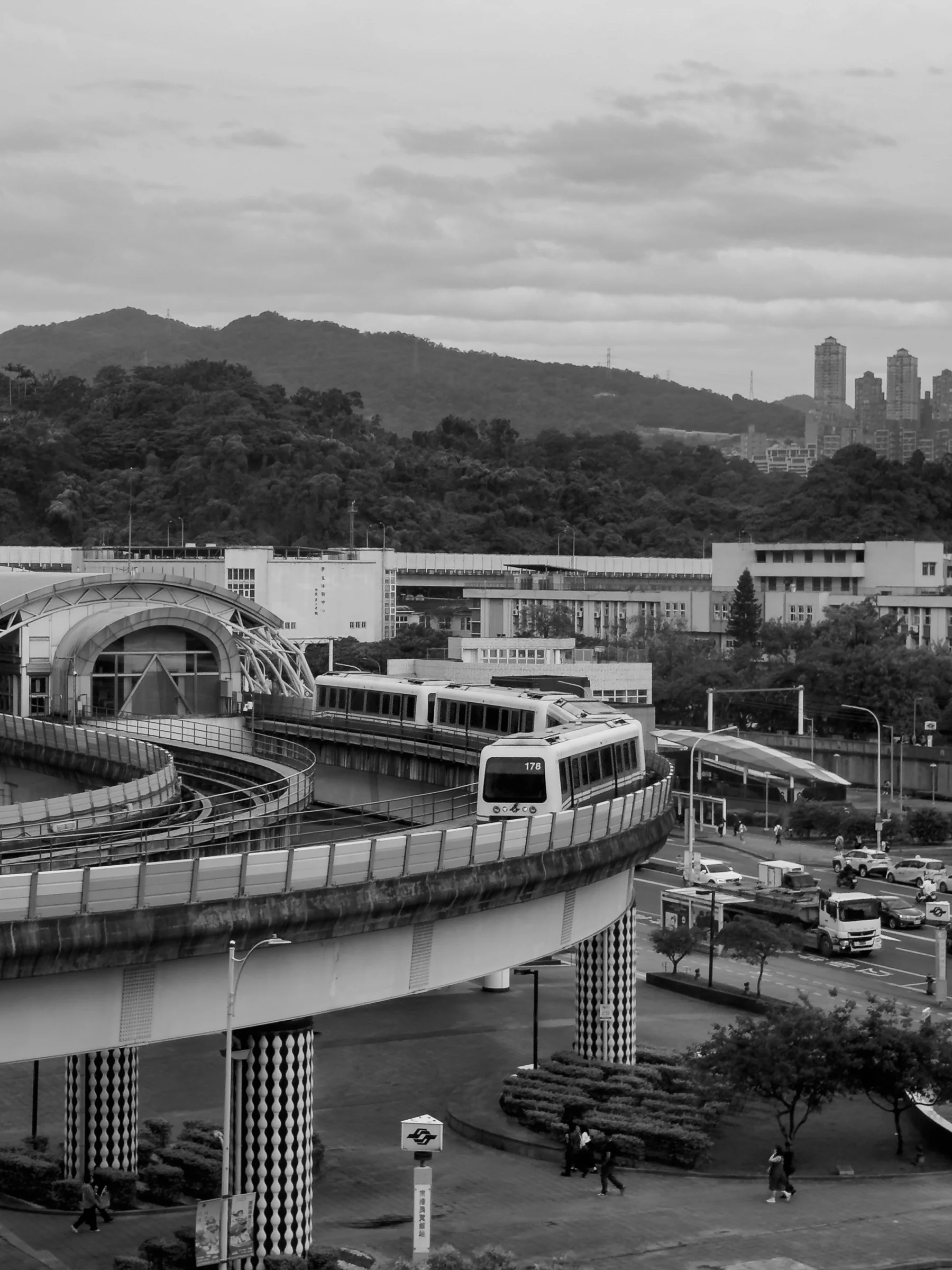 Curving elevated tracks pass through buildings beneath hills and a cloudy sky in black and white.