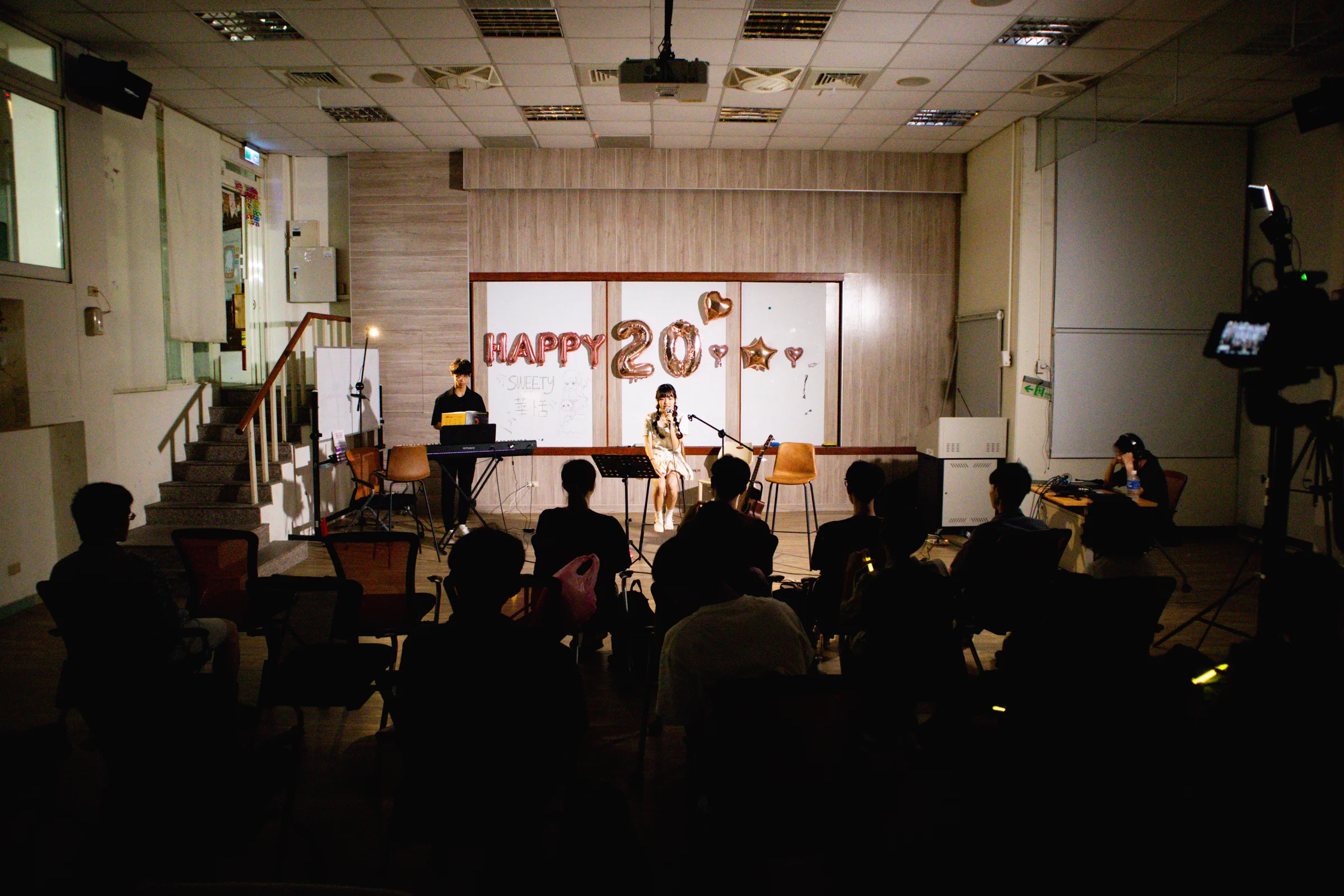 An audience watches a performance in a small room with a bright backdrop.