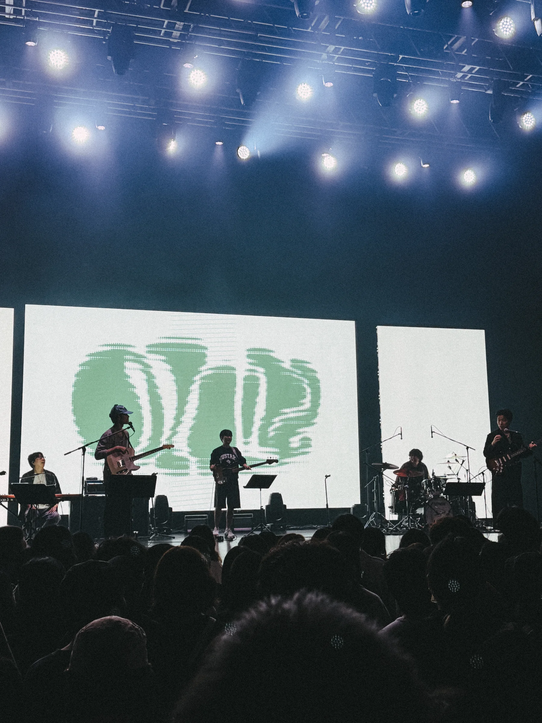 Silhouetted performers stand beneath bright overhead lights in front of a large screen.
