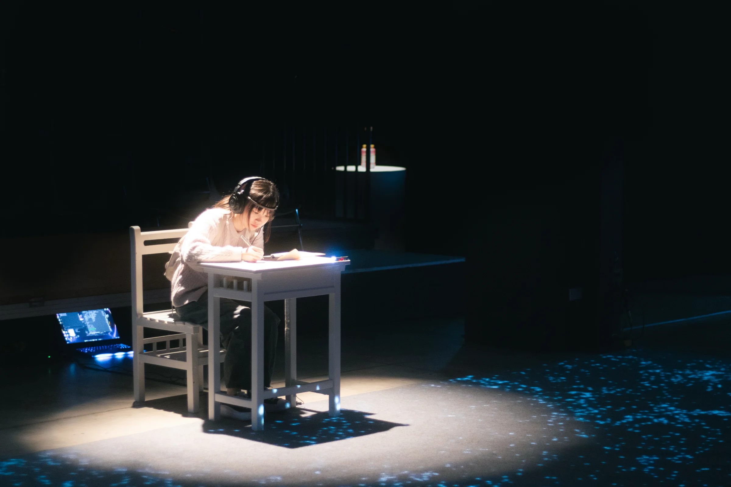 A lone performer sits at a desk in a bright pool of light on a dark stage.