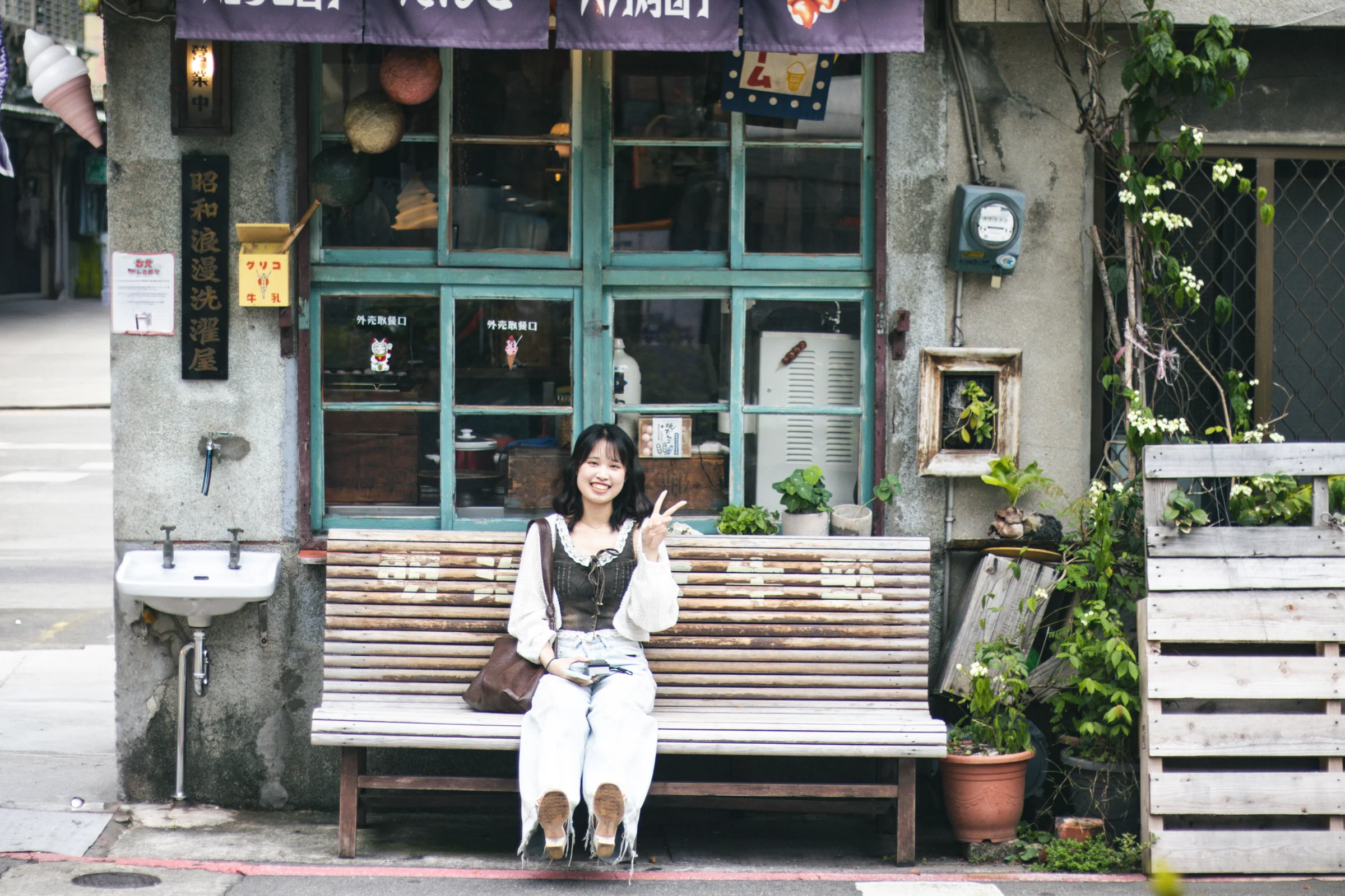 A person sits on a wooden bench in front of a shop with green window frames.