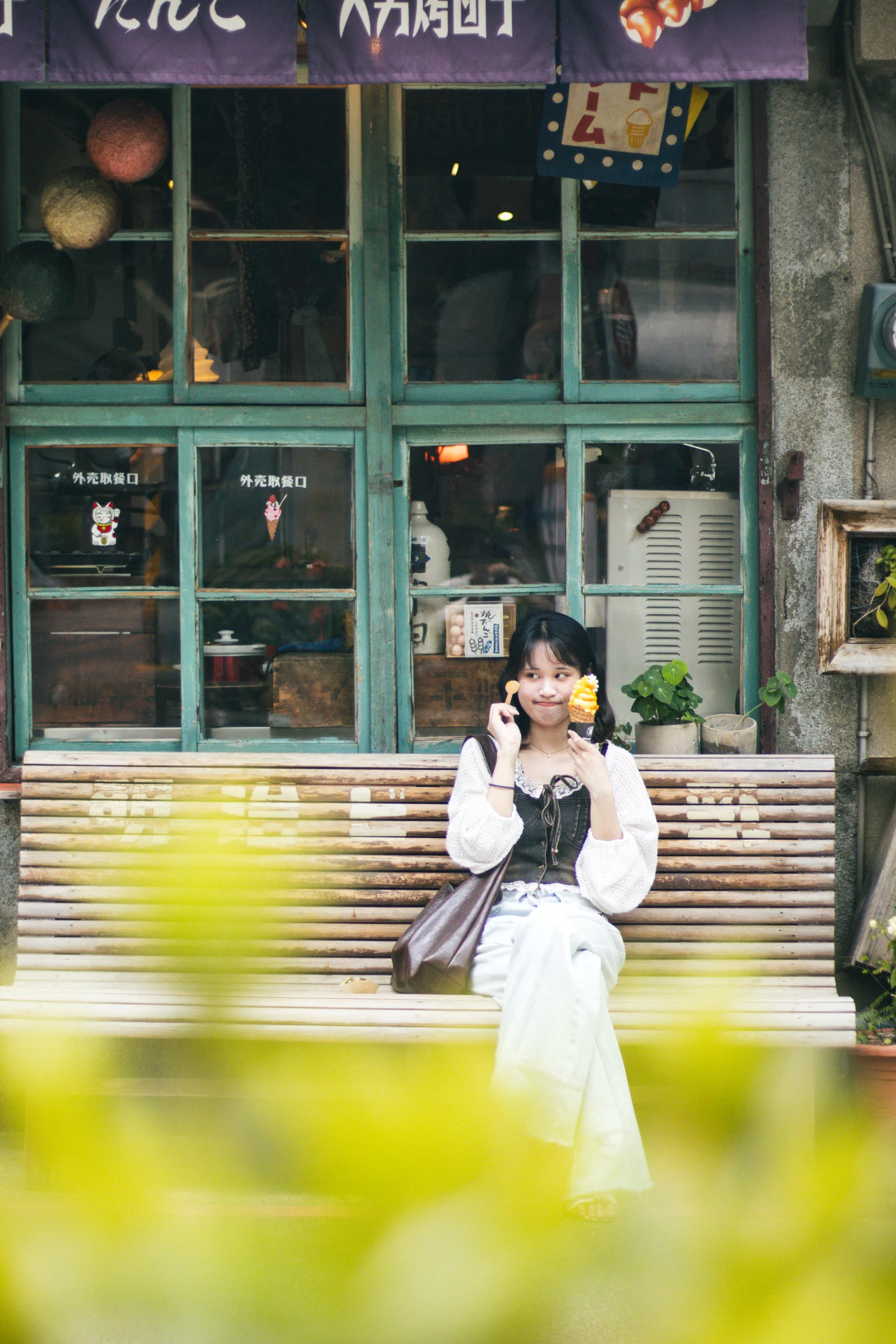 Leaves blur across the foreground as a person sits outside a green-fronted shop.