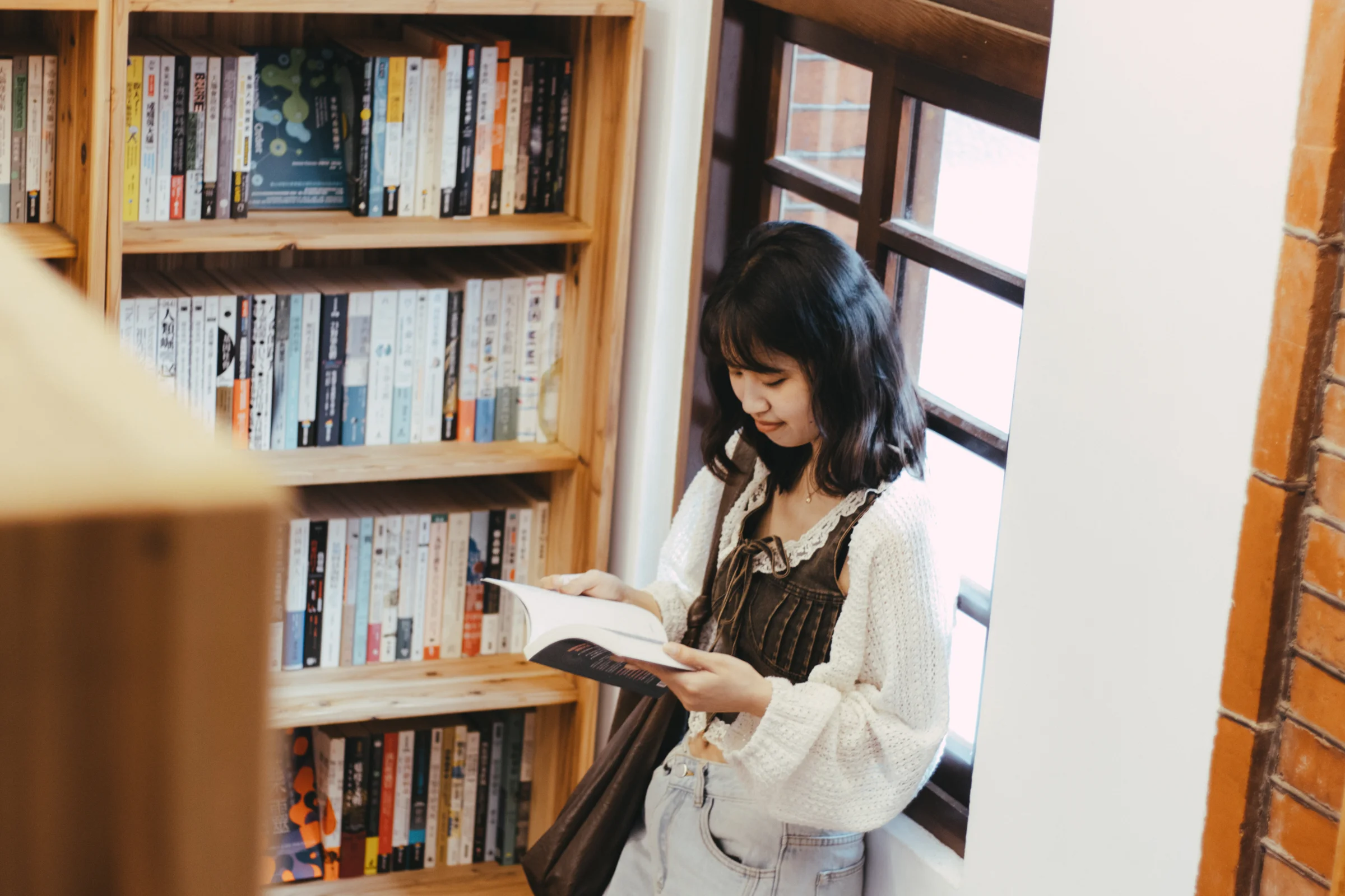 A person looks through a book beside wooden bookshelves in warm light.