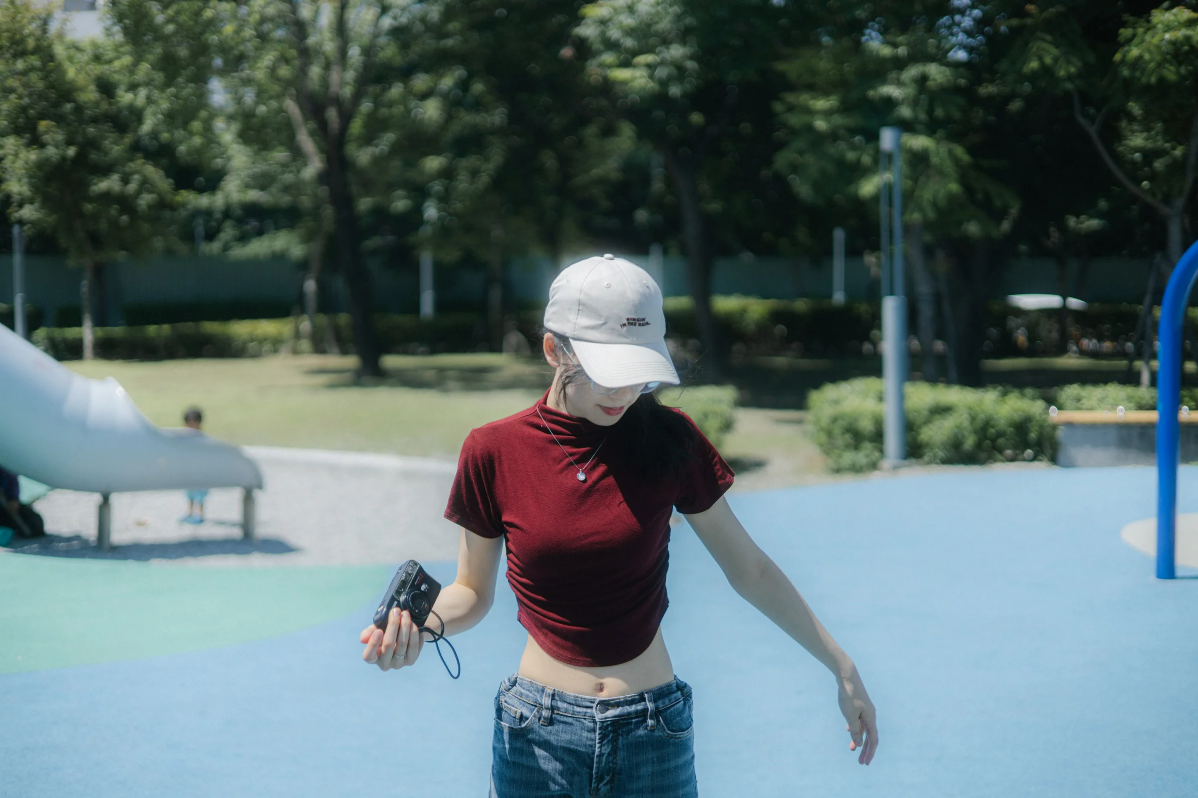 A person in a white cap and red top looks down while standing in a sunny park.