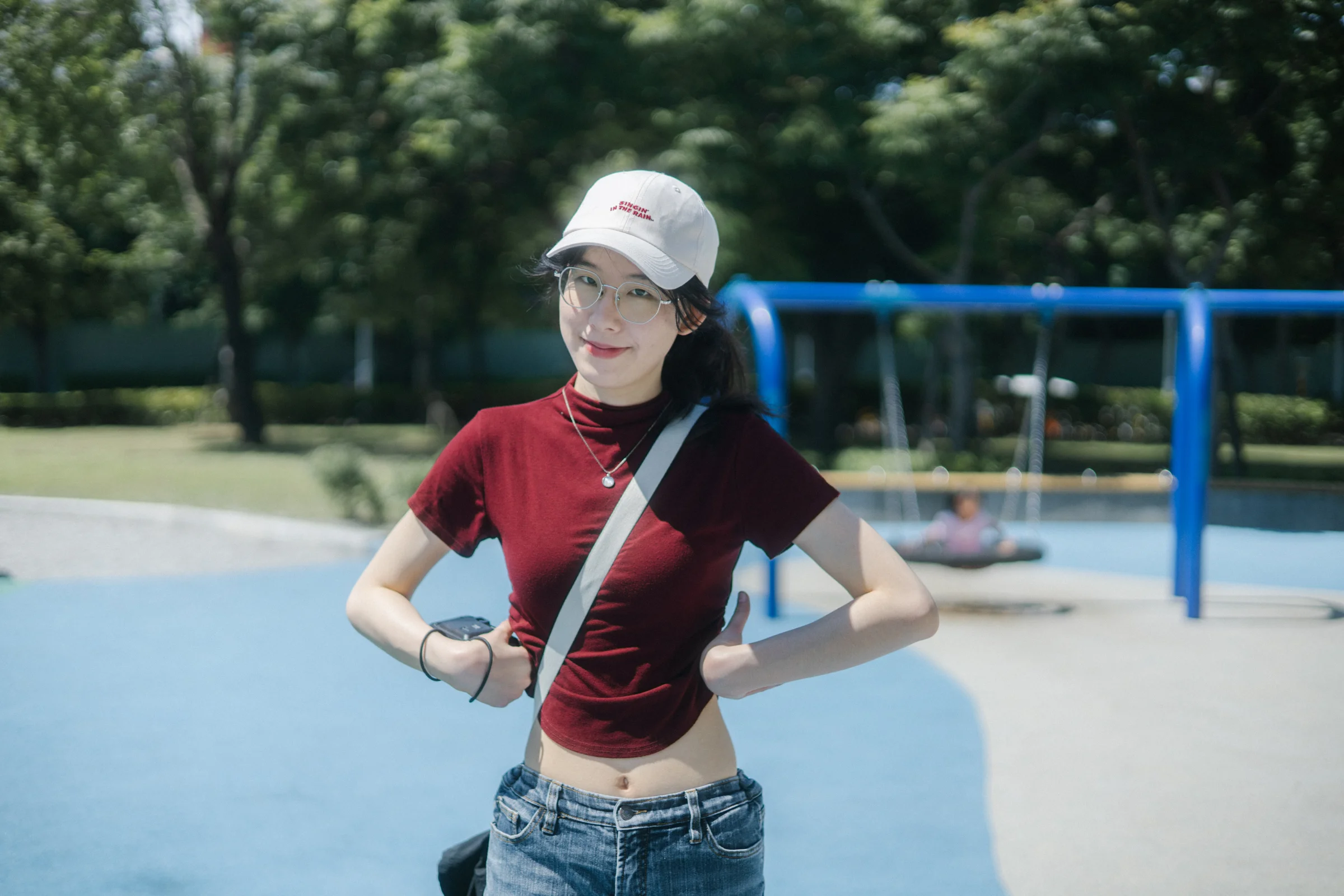 A person in a white cap and red top stands with their hands on their hips in a park.