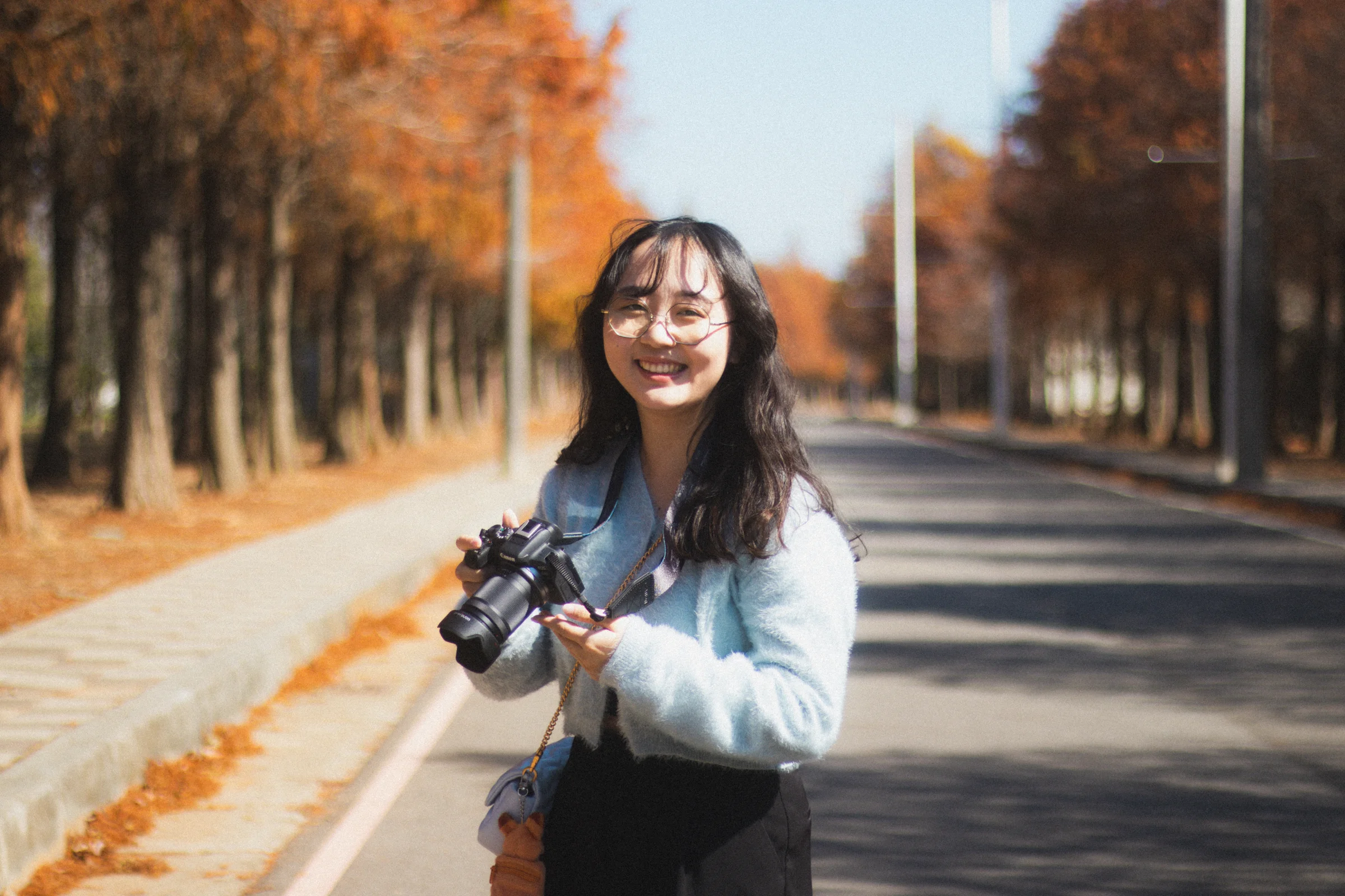 A person holding a camera smiles on a tree-lined road in autumn sunshine.