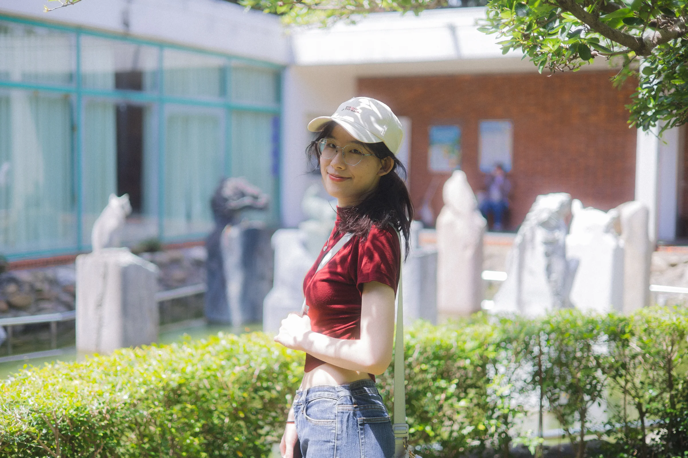 A person in a white cap and red top stands beside a hedge and a brick building.