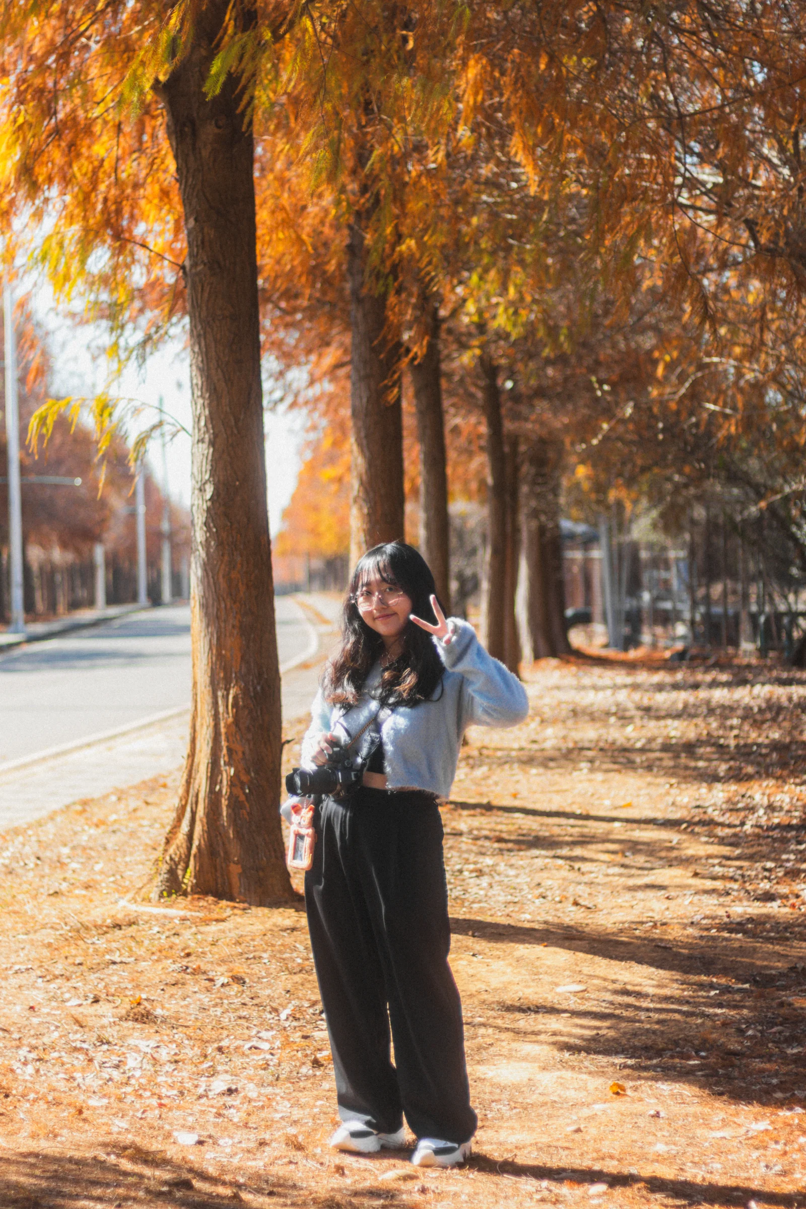 A person stands beside a tree trunk amid orange foliage and fallen leaves.