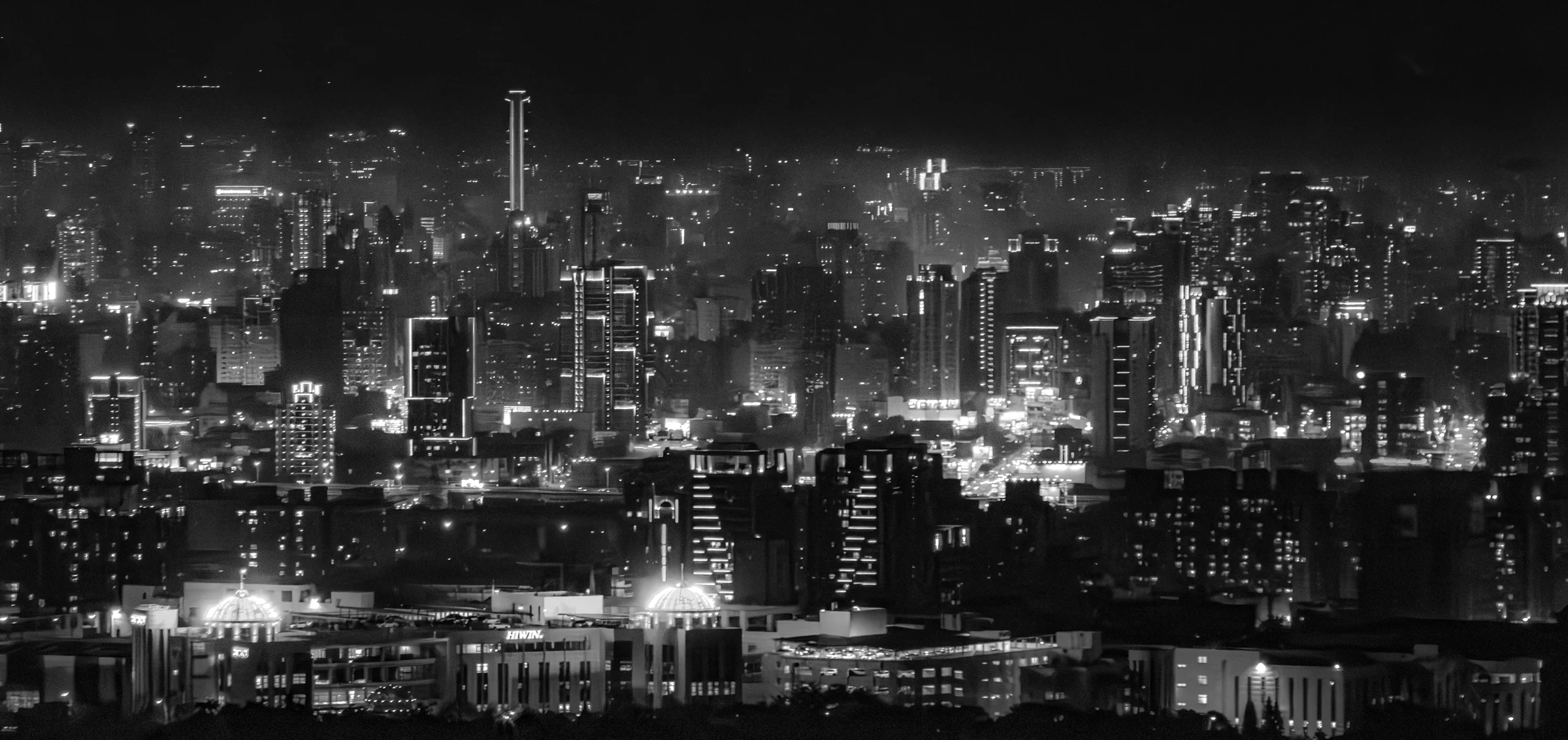 A dense city skyline glows with lights beneath a dark sky in black and white.