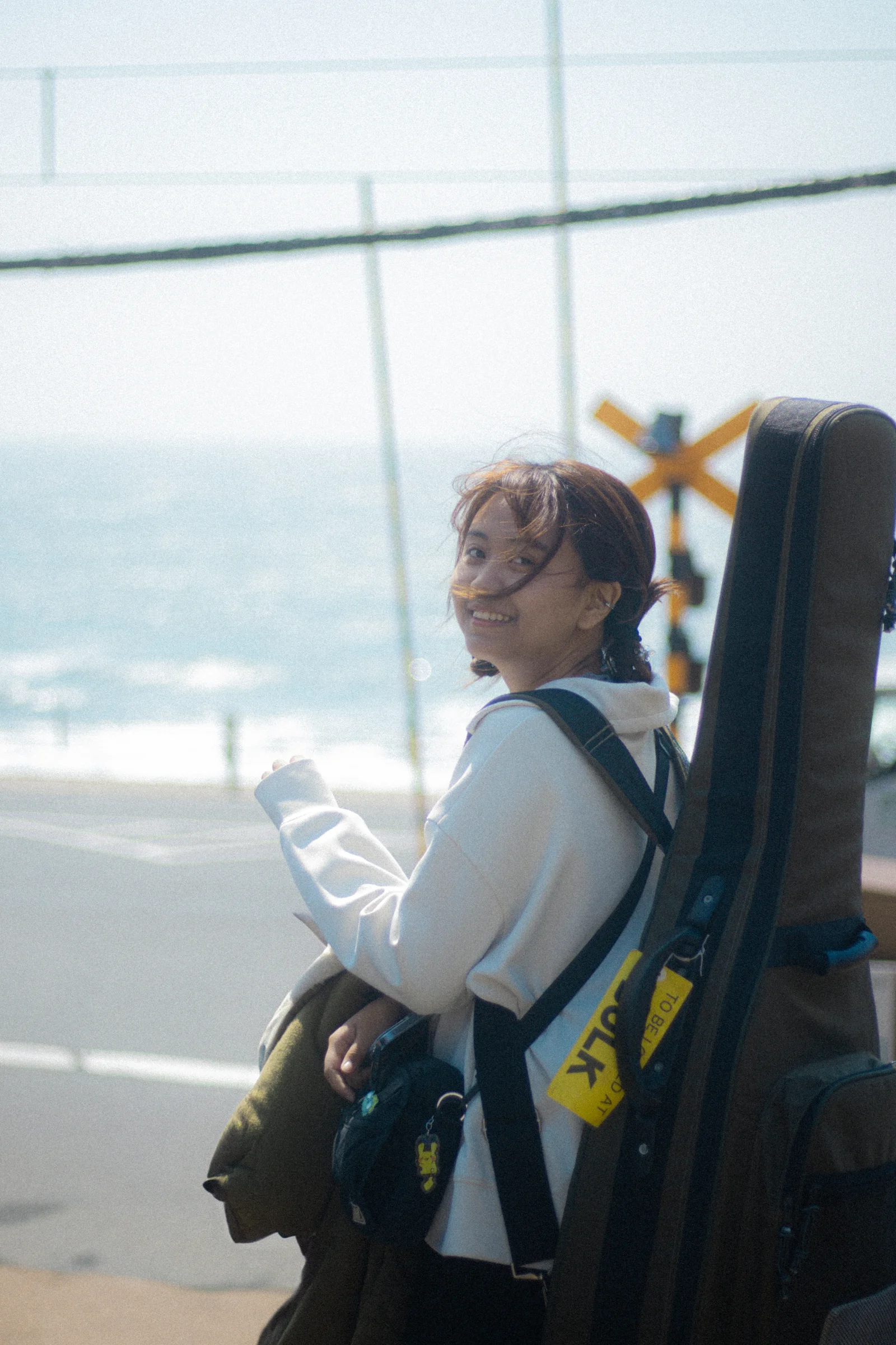 A person carrying a guitar case stands beside a coastal railway crossing.