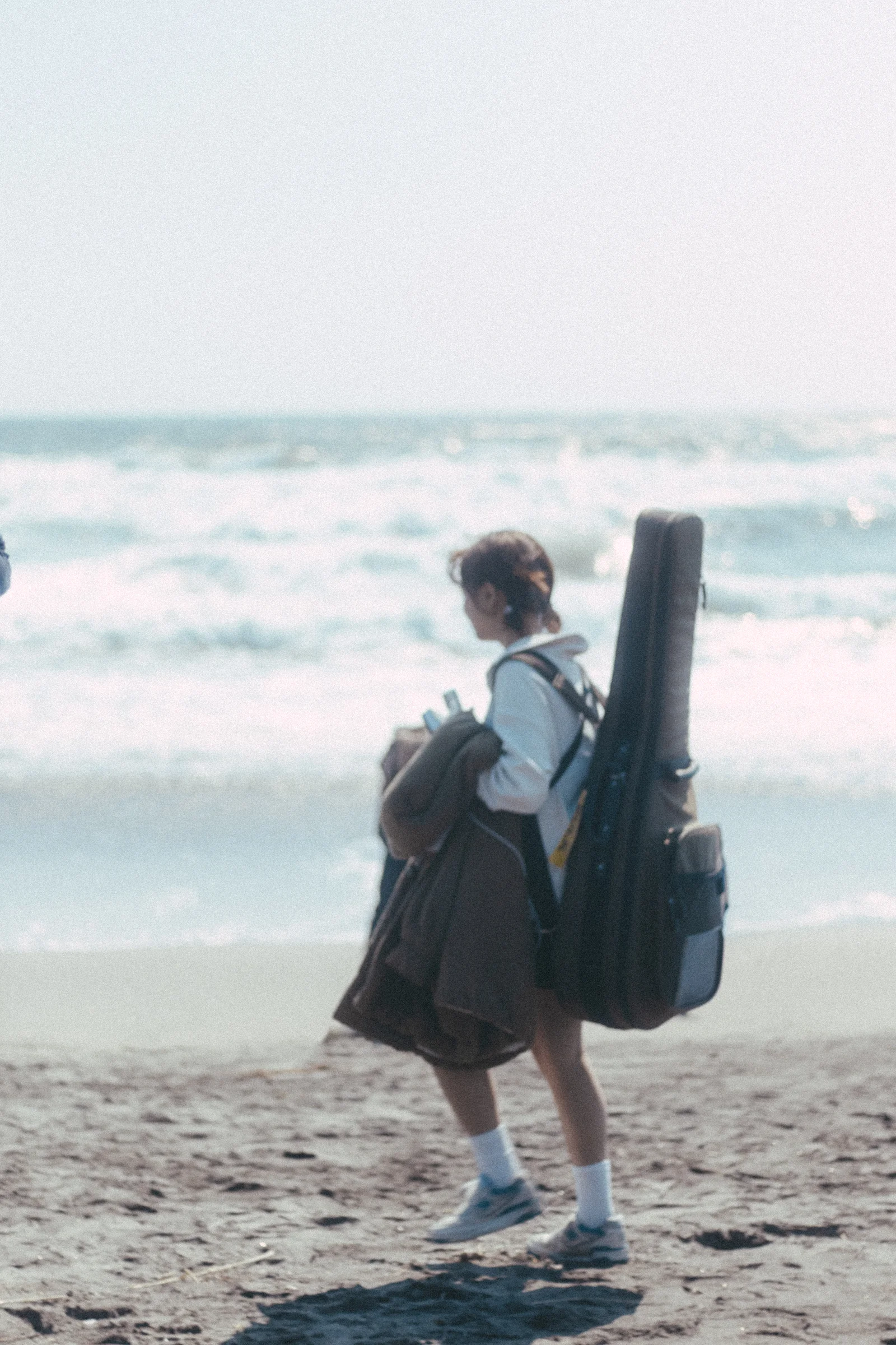 A person with a guitar case stands on the beach facing the waves.