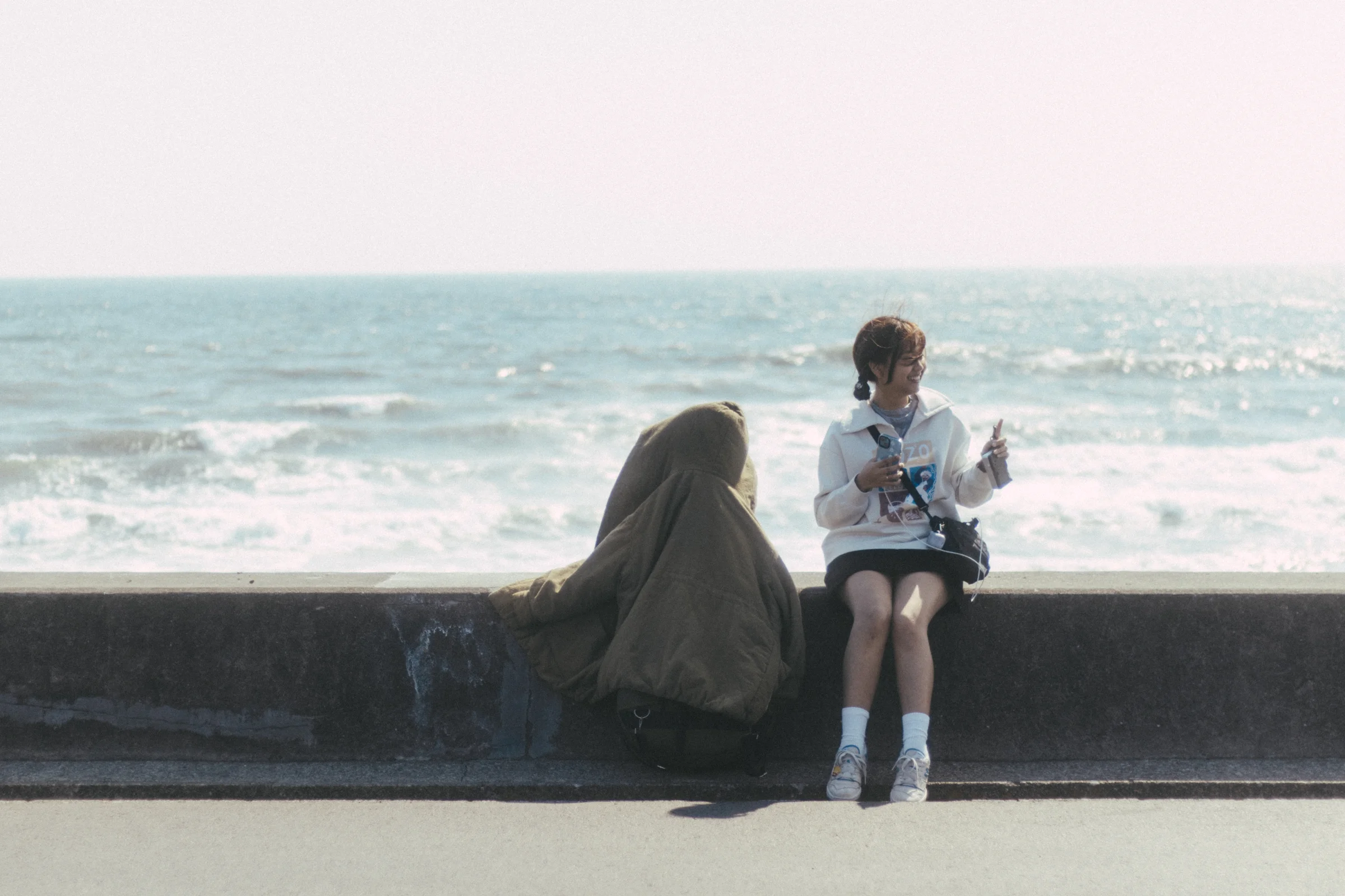 A person sits on a seawall beside a guitar case with waves behind them.