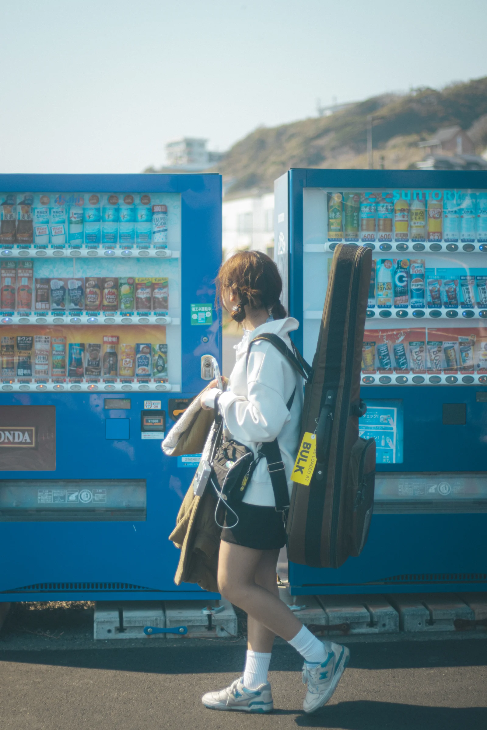 A person carrying a guitar case walks past two blue vending machines beneath a sunlit hillside.