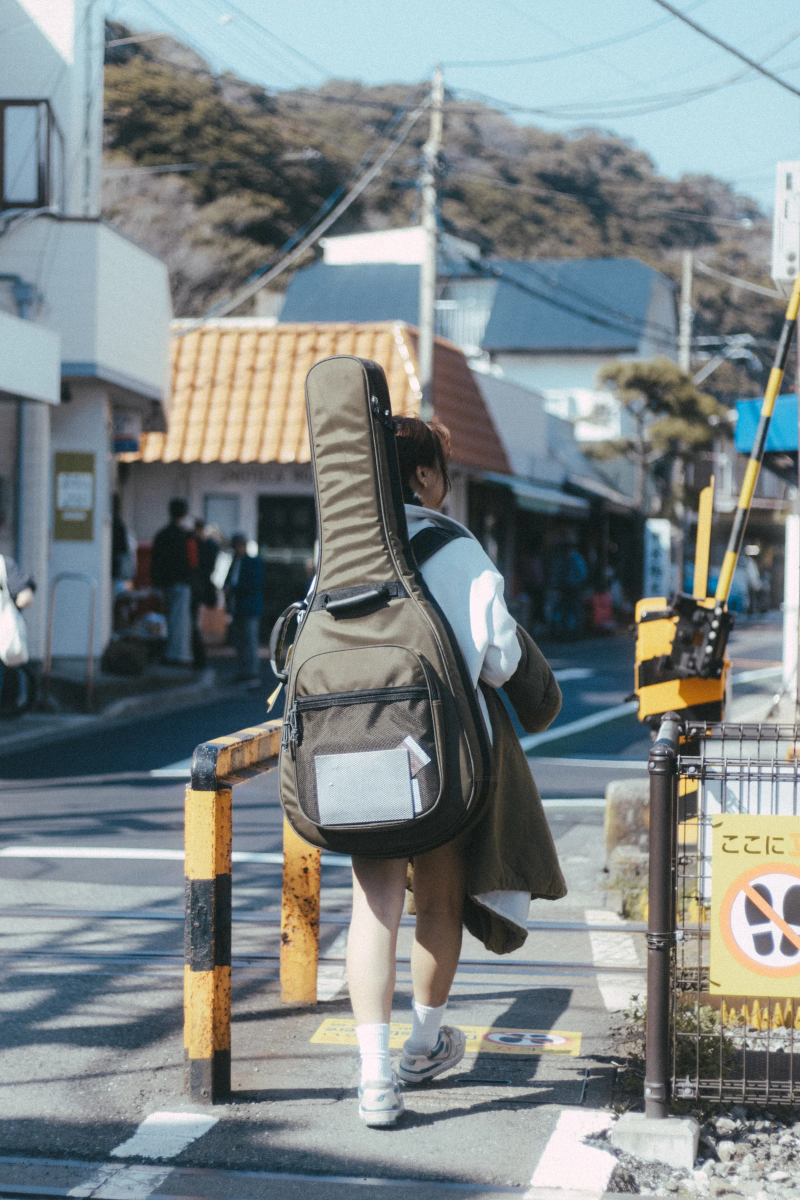 A person carrying a guitar case walks across railway tracks between small buildings.