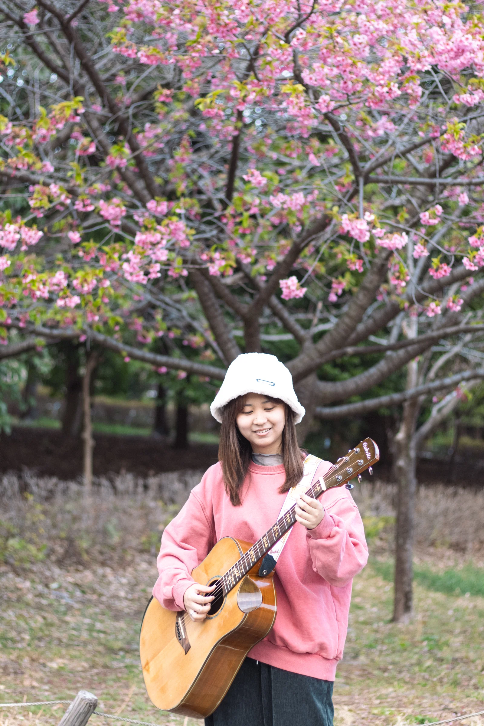 A person in a white cap and pink top holds a guitar beneath flowering trees.