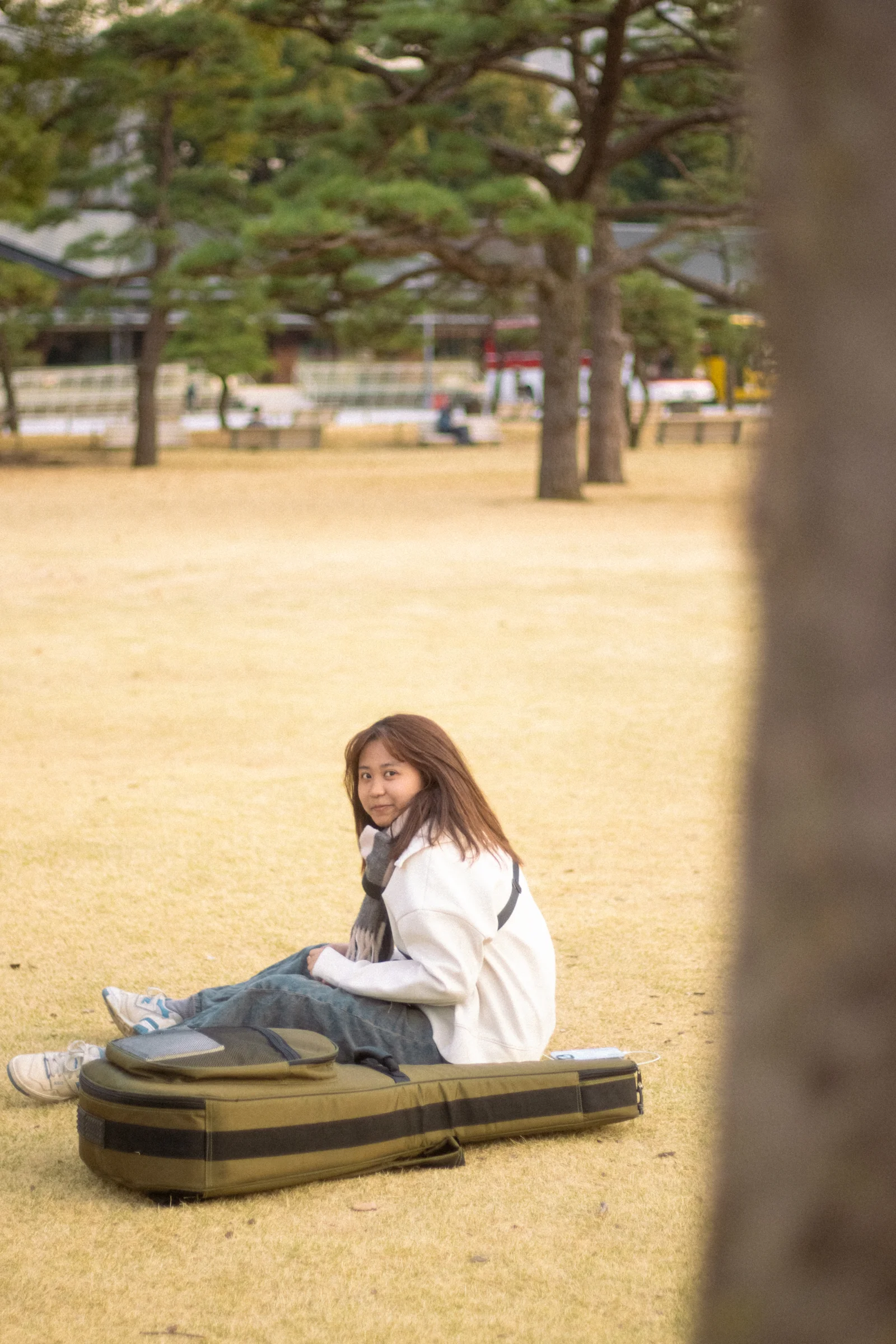 A person sits on a lawn beside a guitar case among park trees.