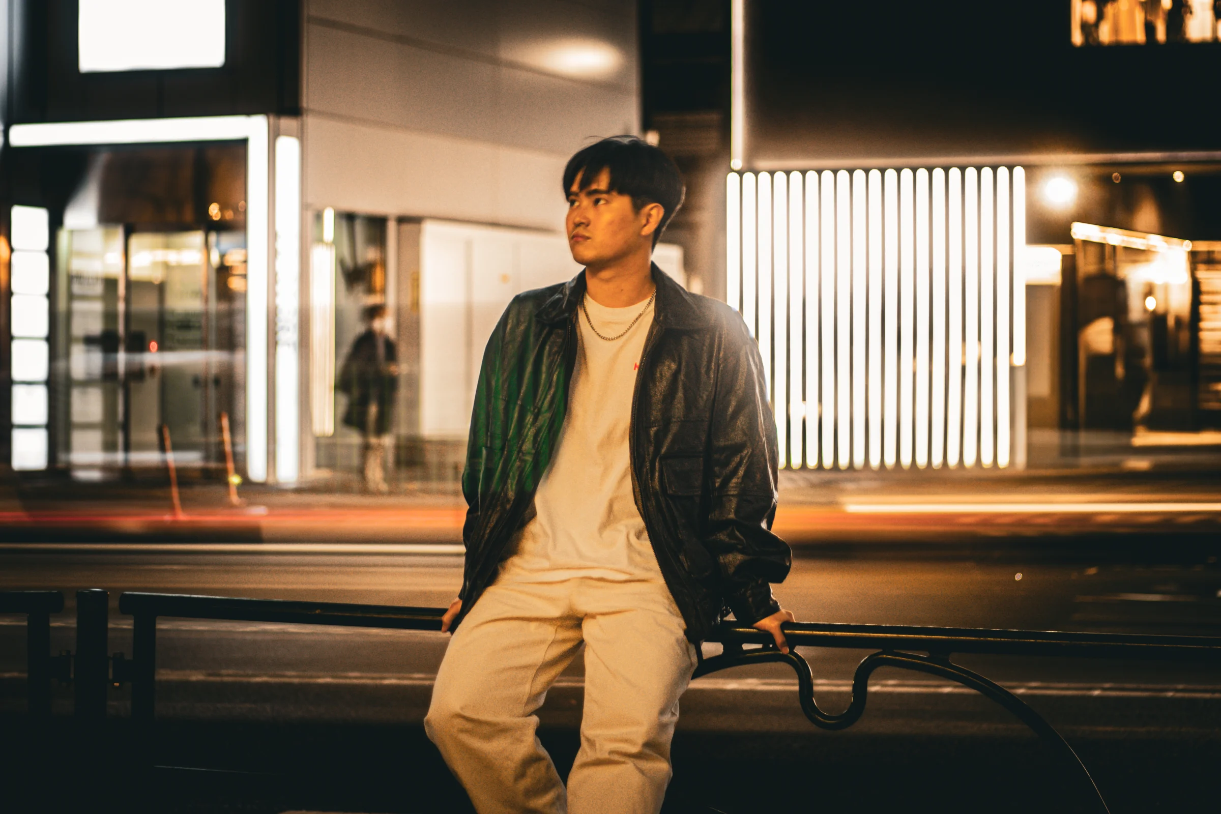 A person sits beside a lit storefront on a city street at night.