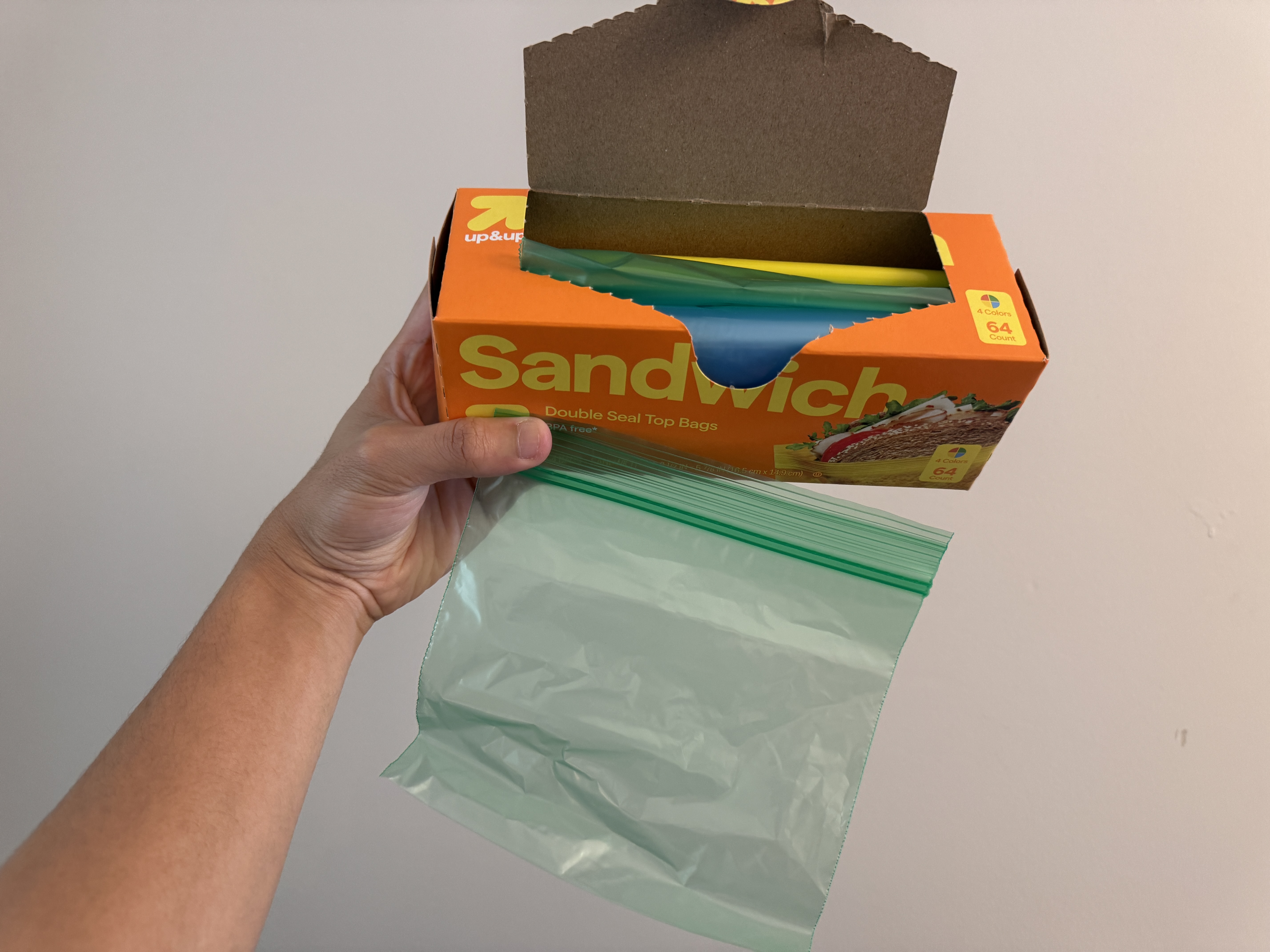 A hand holding an open box of colorful zip-top bags with one transparent bag pulled out.