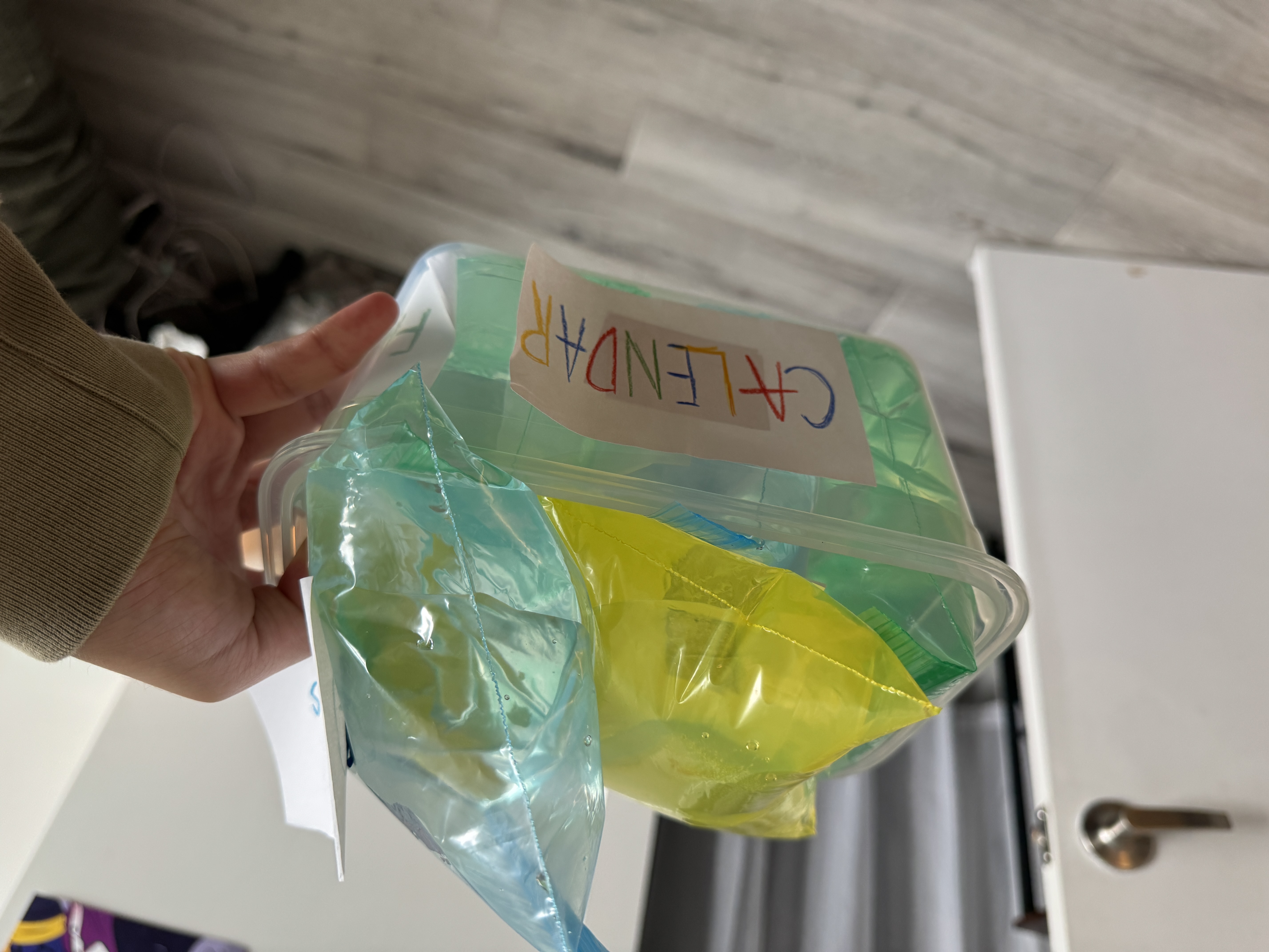 A hand lifting a transparent container labeled CALENDAR, filled with blue, green, and yellow water bags.