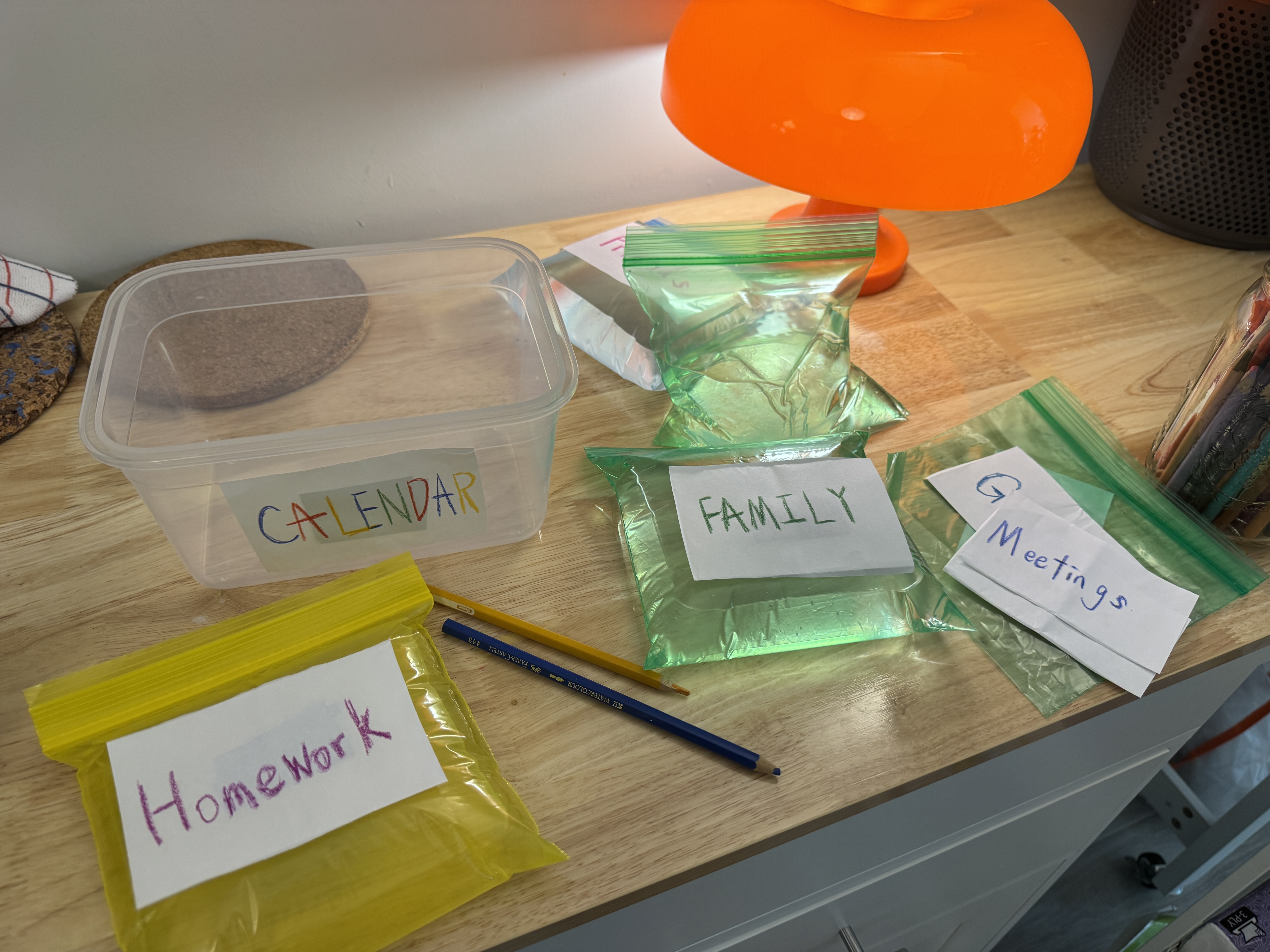 A transparent CALENDAR container beside colorful water-filled activity bags labeled Homework, Family, and Meetings on a wooden table.