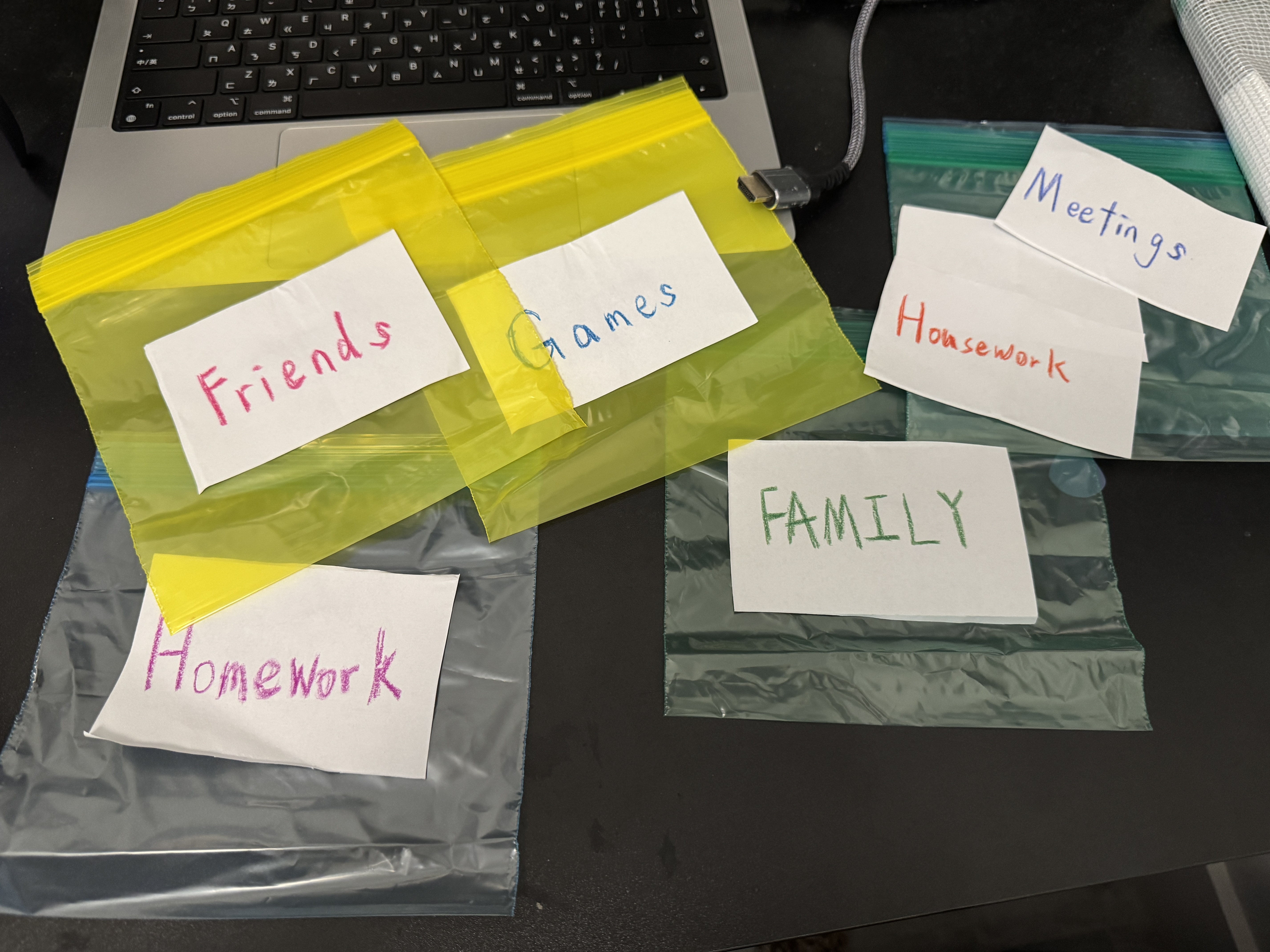 Empty colorful zip-top bags labeled Homework, Friends, Games, Family, Housework, and Meetings arranged beside a laptop.